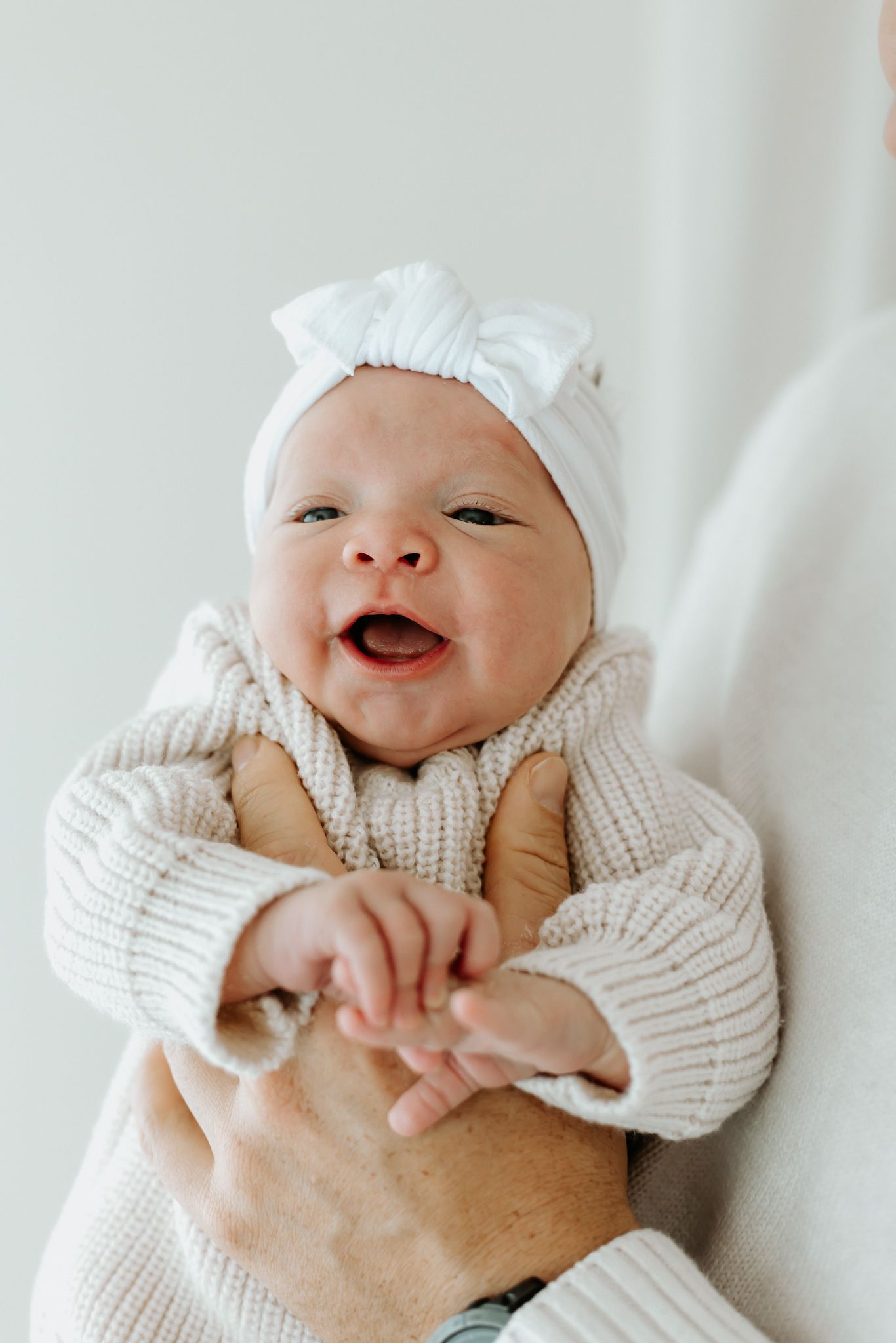 Adorable newborn baby smiling in a white bow headband and beige knit sweater, cradled in loving arms.