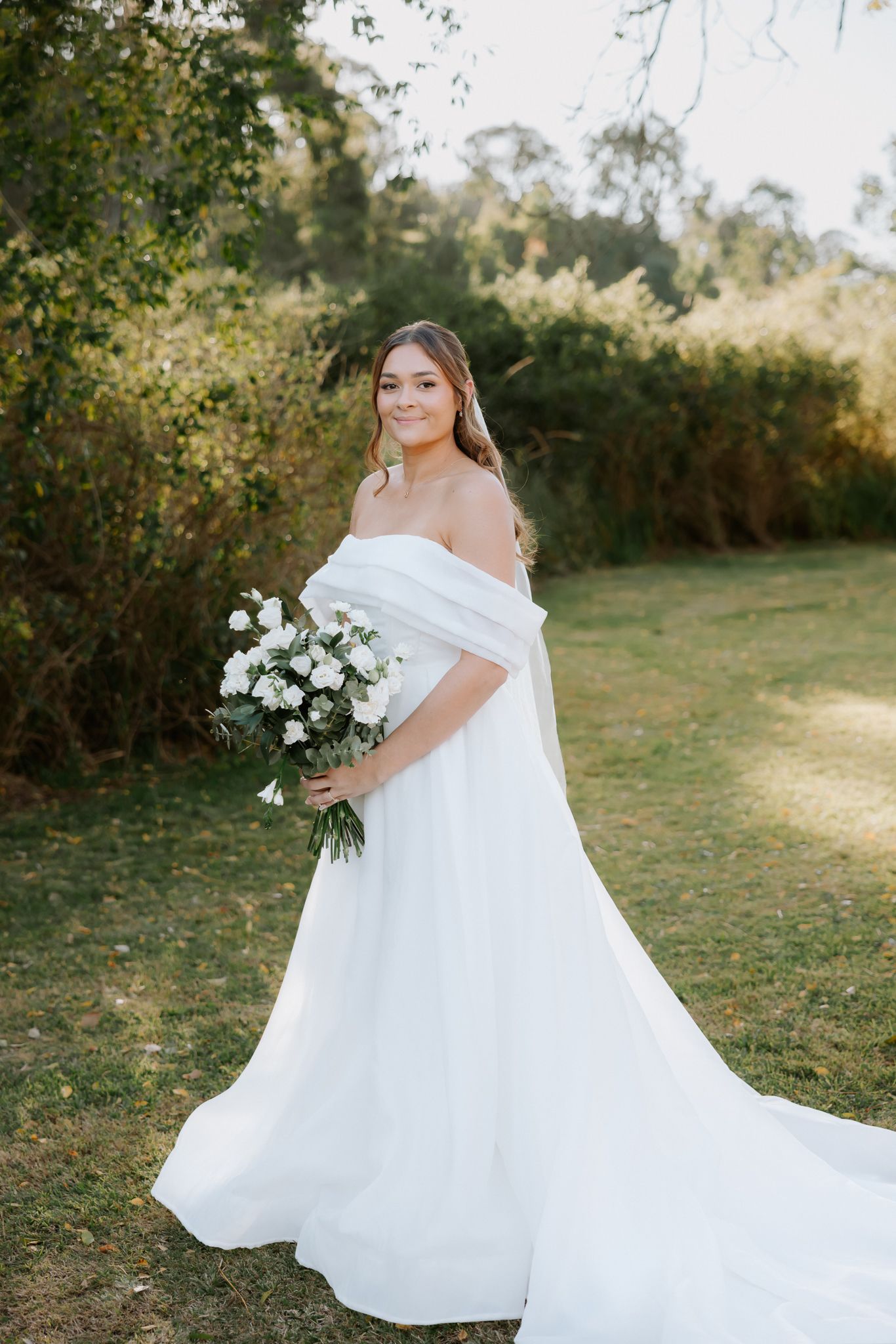 Bride in off-shoulder white gown holding white floral bouquet, standing outdoors in lush green garden setting.