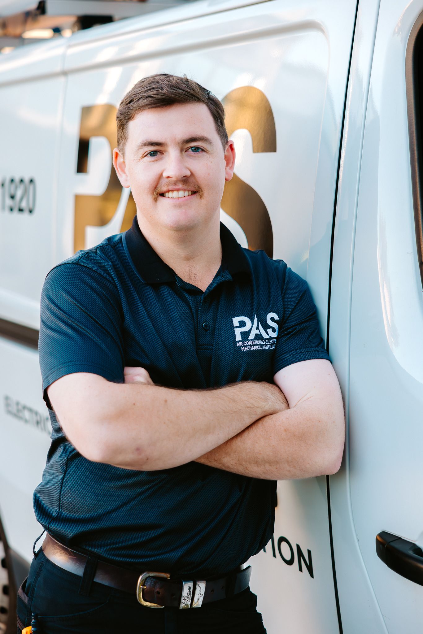Smiling PAS Air Conditioning technician in navy polo shirt leaning against company van with arms crossed.