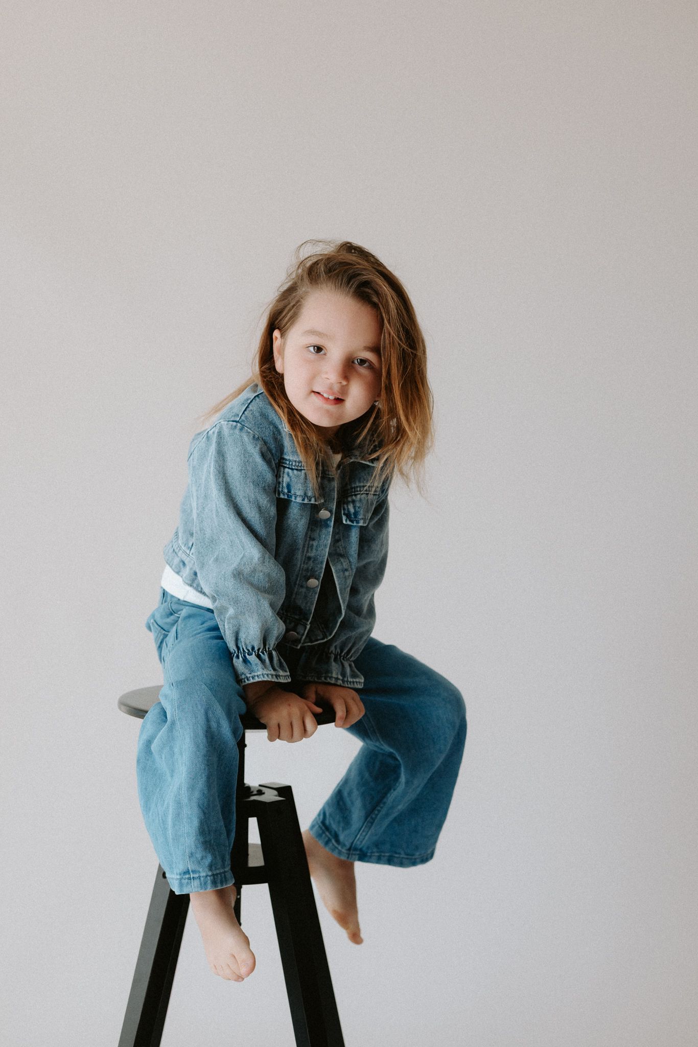 Young girl in denim jacket and jeans sitting barefoot on a stool against a white background.
