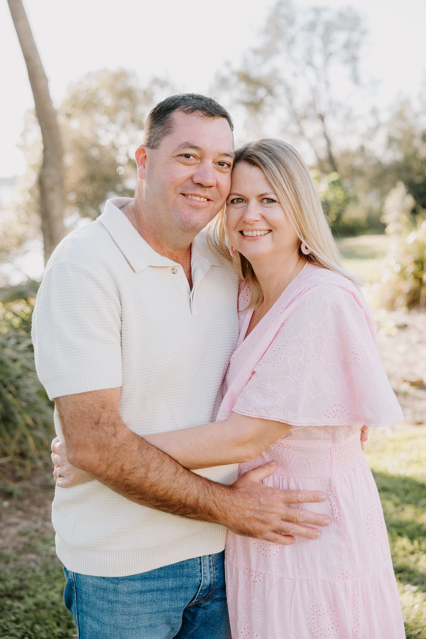 A smiling couple embracing outdoors in a sunny park, man in cream polo and jeans, woman in pink dress.