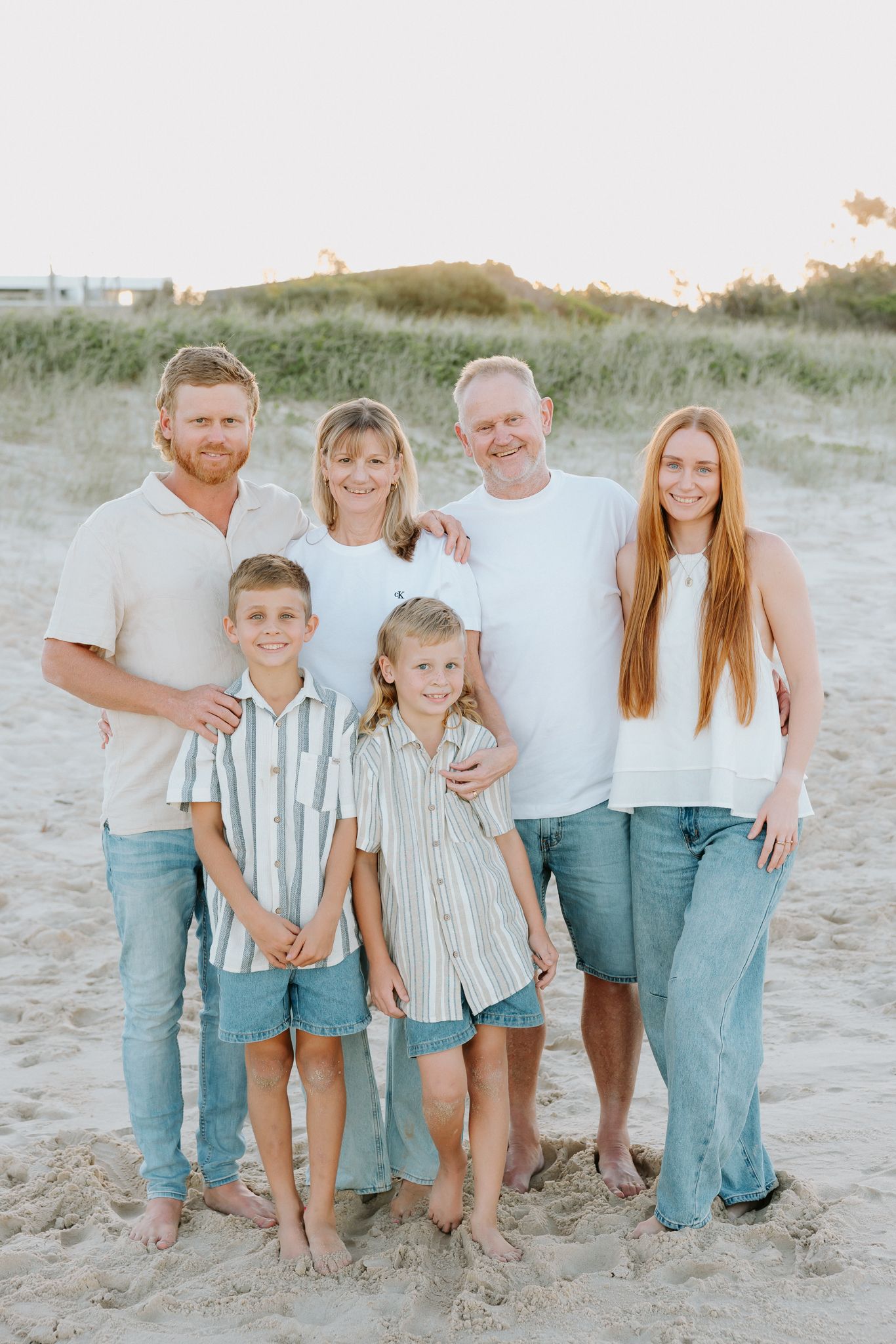 Family of six posing together on a sandy beach at golden hour, wearing coordinated casual outfits of white tops and jeans.