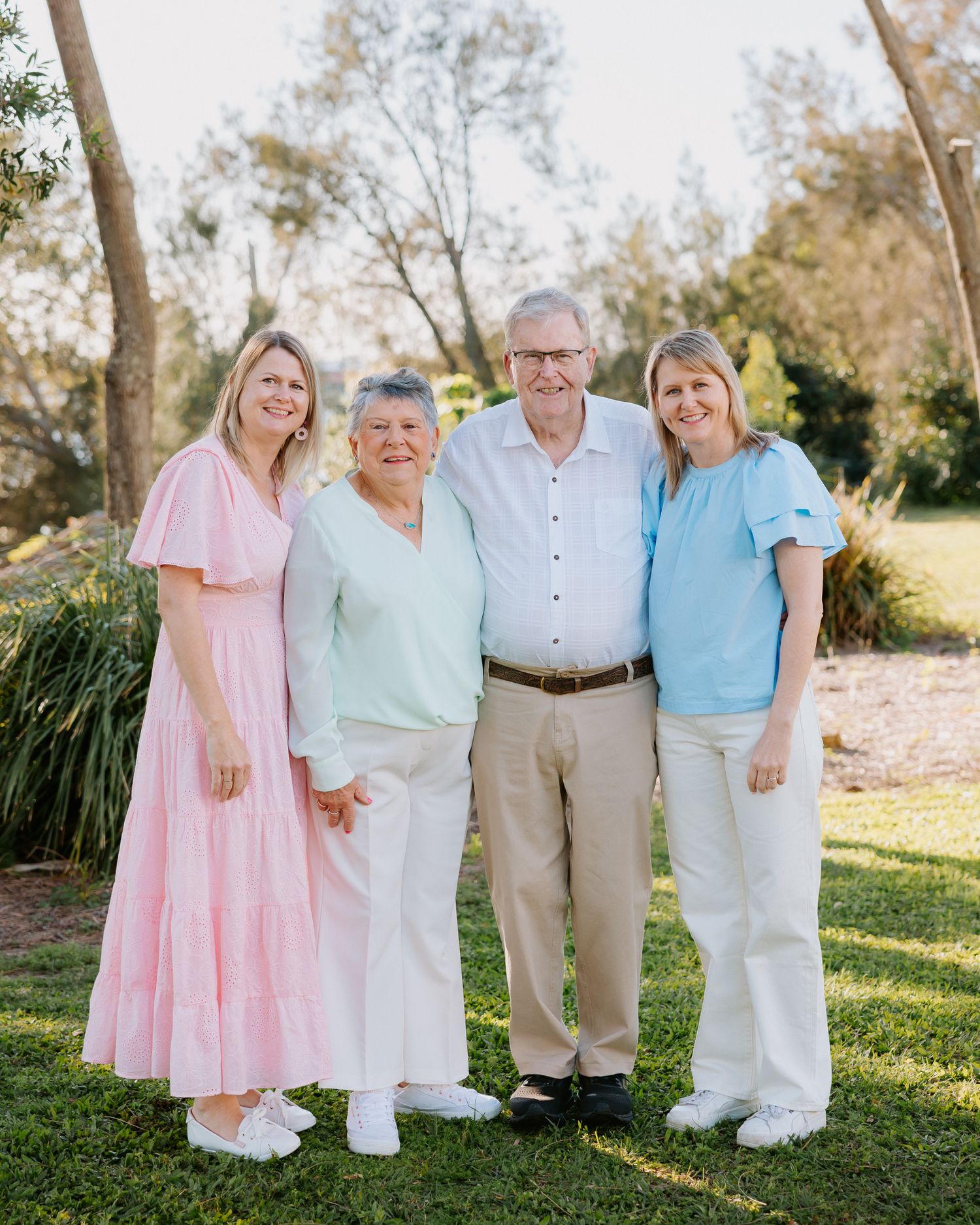 Four smiling family members pose together outdoors on green grass surrounded by trees in a warm, sunny setting.