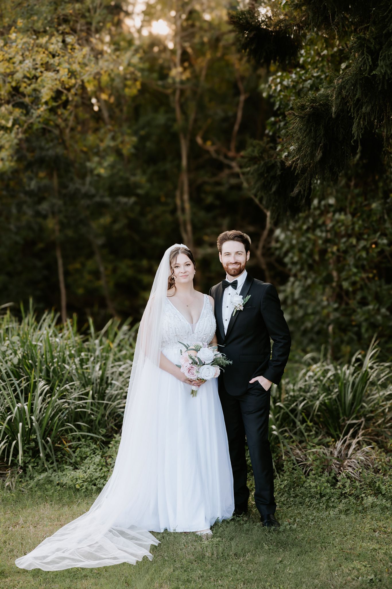 Bride in white gown with veil holding bouquet stands beside groom in black tuxedo in lush garden setting.