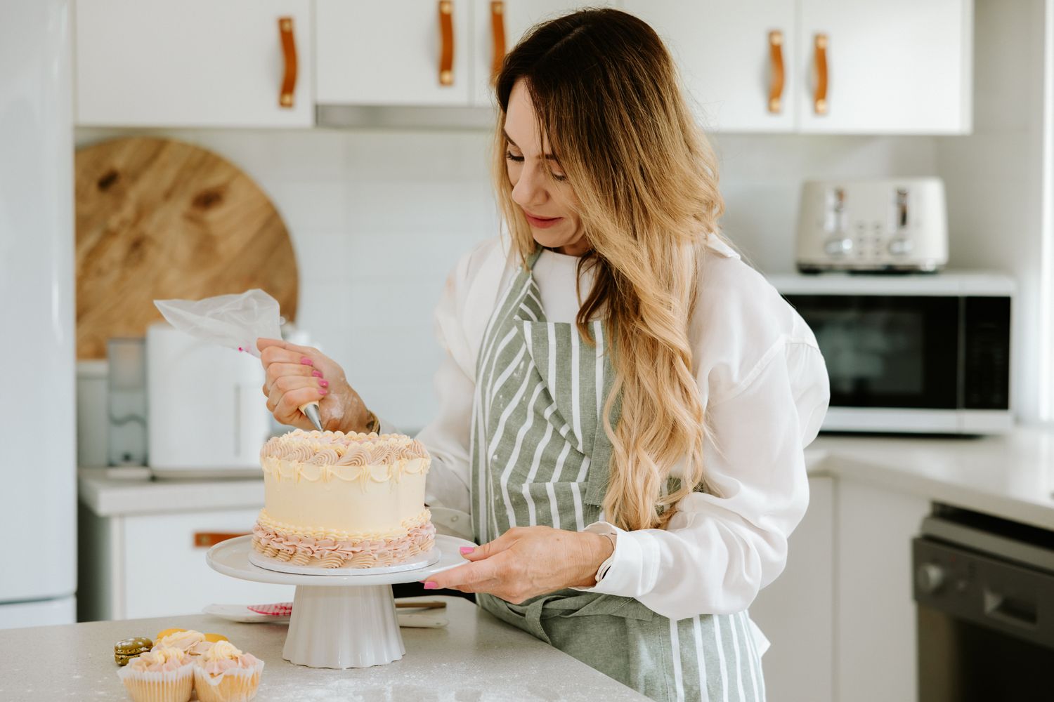 Woman in striped apron decorating a layered frosted cake on a white cake stand in a bright modern kitchen.