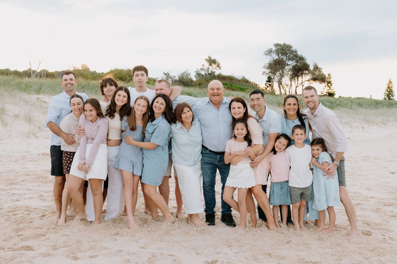 Large family group portrait on sandy beach, smiling multi-generational family in coordinated pastel outfits with dunes behind.