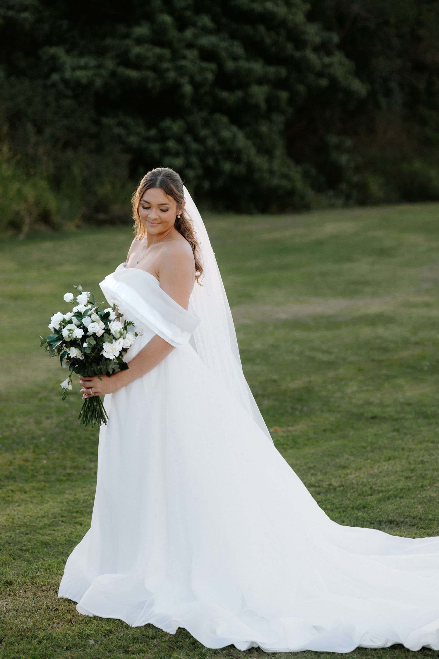 Bride in white off-shoulder gown with veil holding floral bouquet on green lawn.