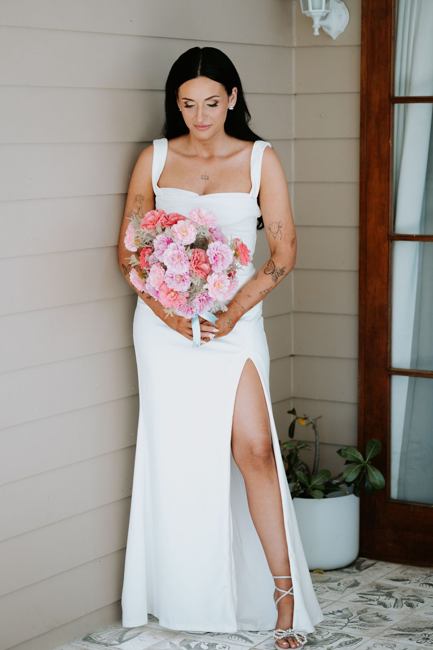 Bride in elegant white dress with slit holding pink bouquet against neutral wall.