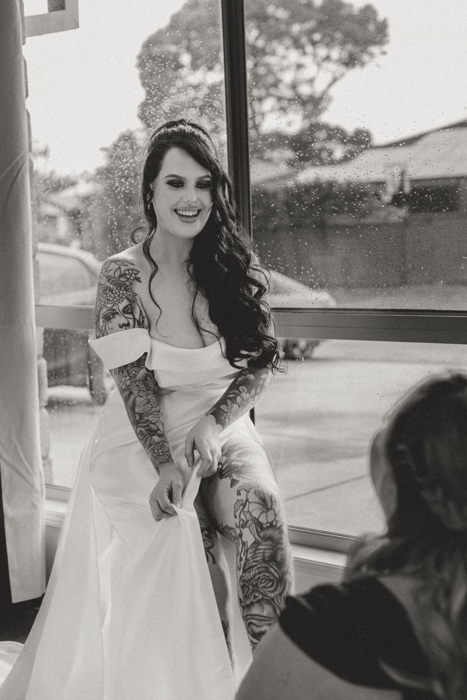 Black and white portrait through window showing bride in elegant lace dress.