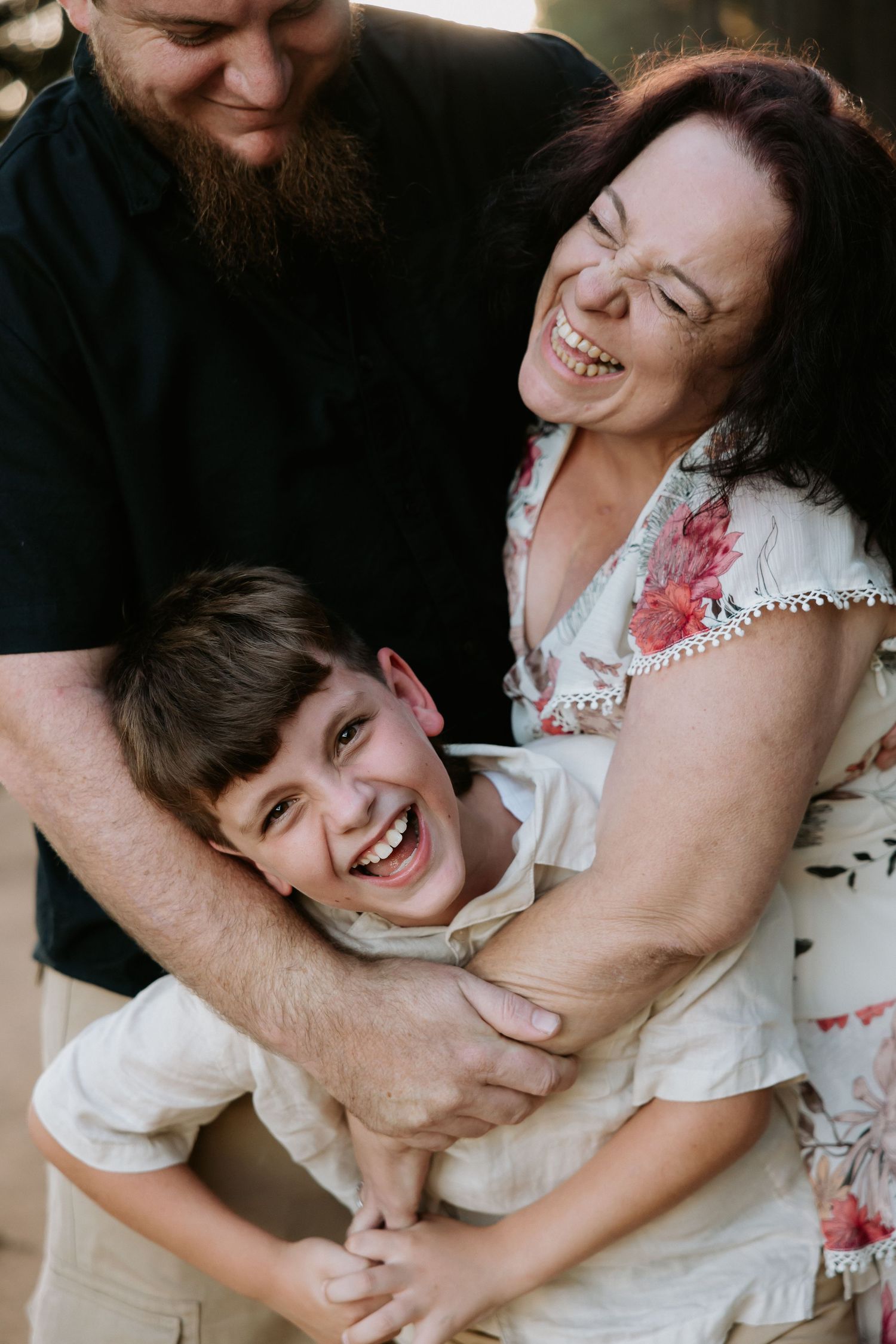 A joyful family moment with a bearded man and woman laughing while hugging a young smiling boy outdoors.