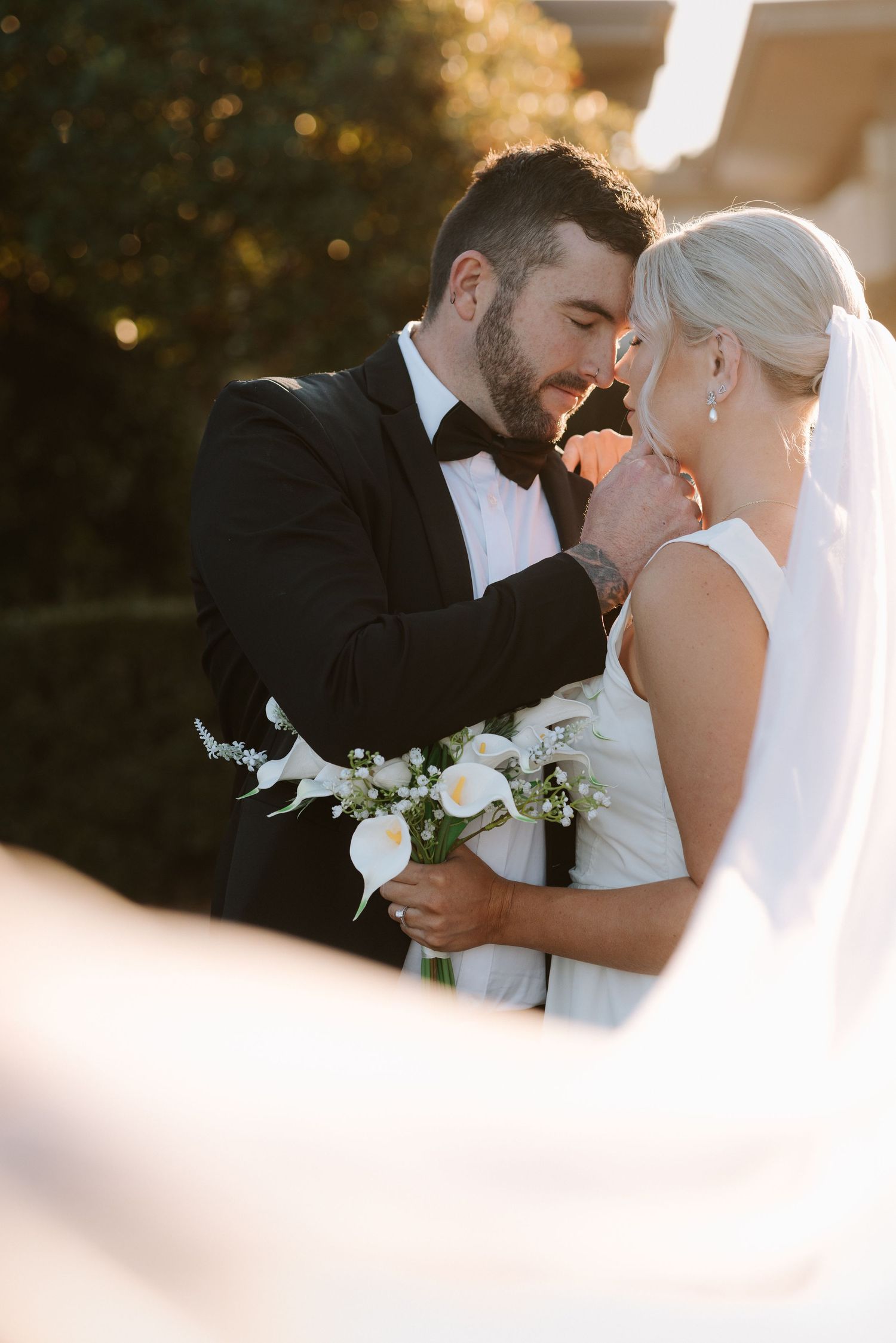 Bride and groom share an intimate moment, groom holding white calla lily bouquet in golden sunset light.