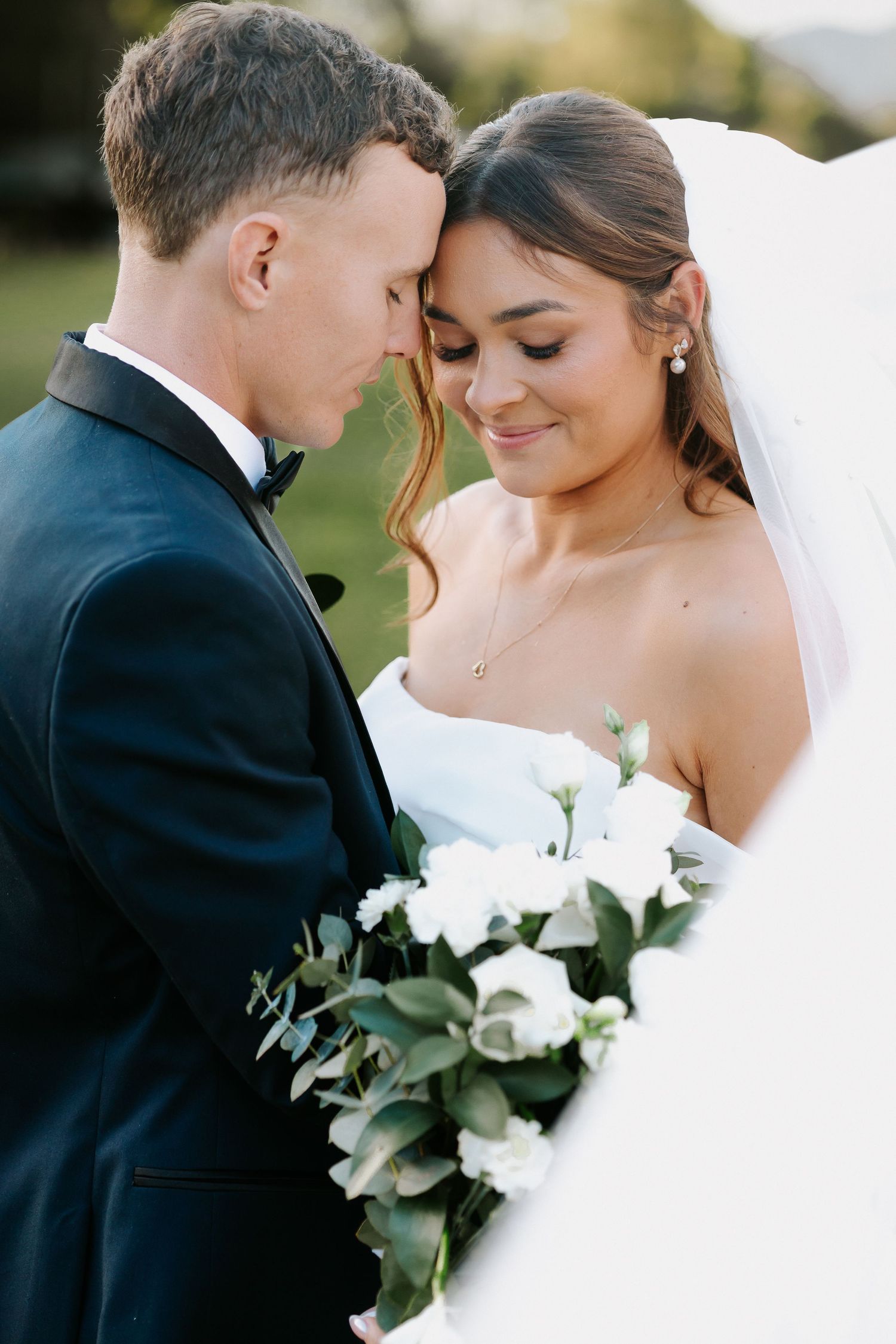 Bride and groom touching foreheads tenderly, bride holding white rose bouquet, both dressed in elegant wedding attire outdoors.