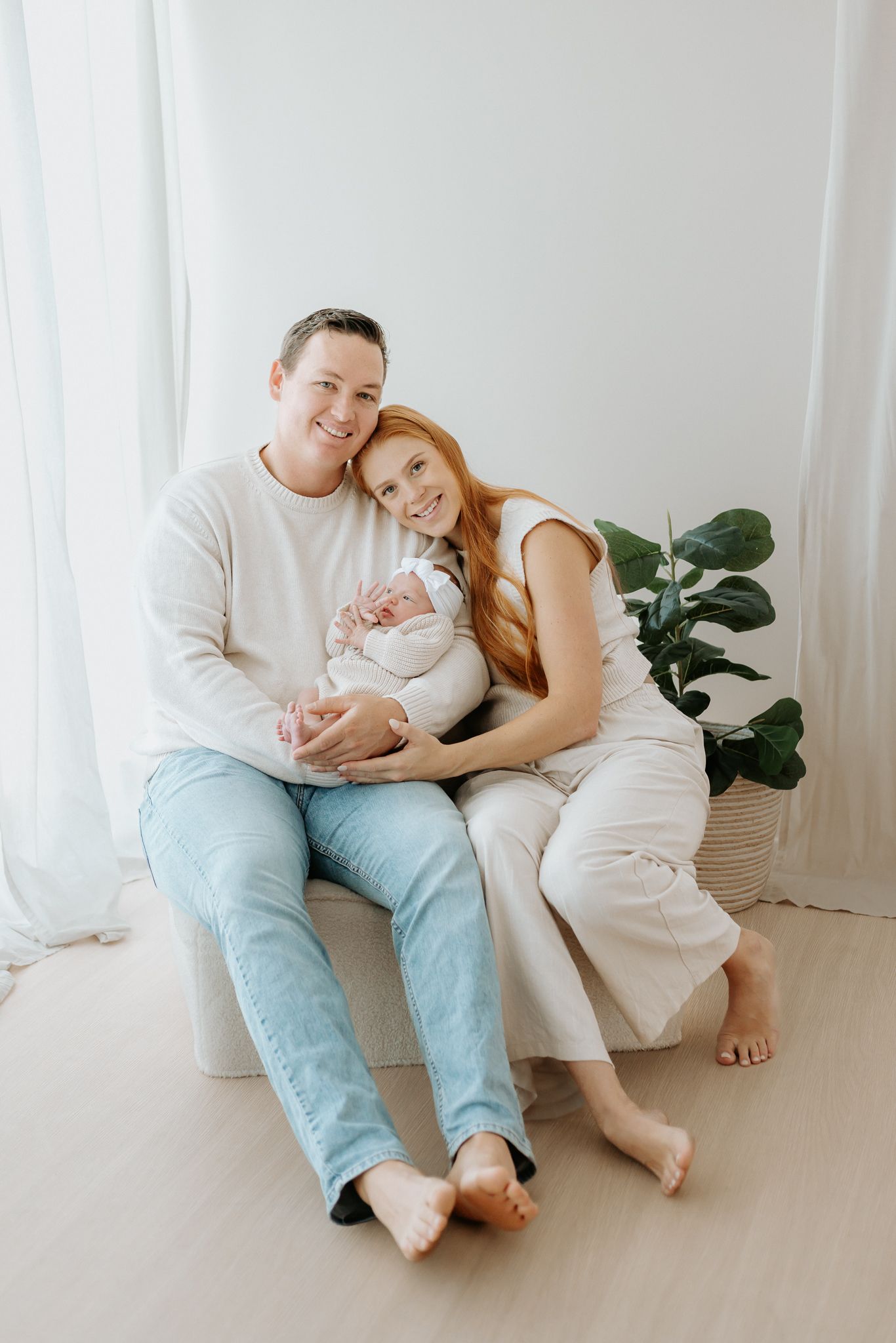 Happy couple sitting barefoot on white couch holding sleeping newborn baby in bright airy studio setting.