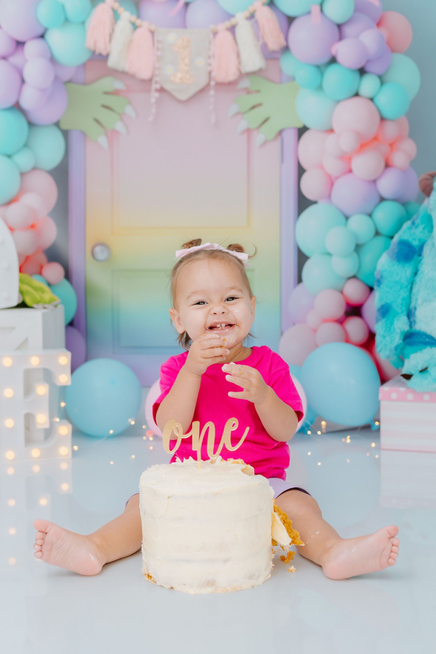 First birthday party setup with pastel balloon arch, white cake, and pink shirt against rainbow gradient backdrop.