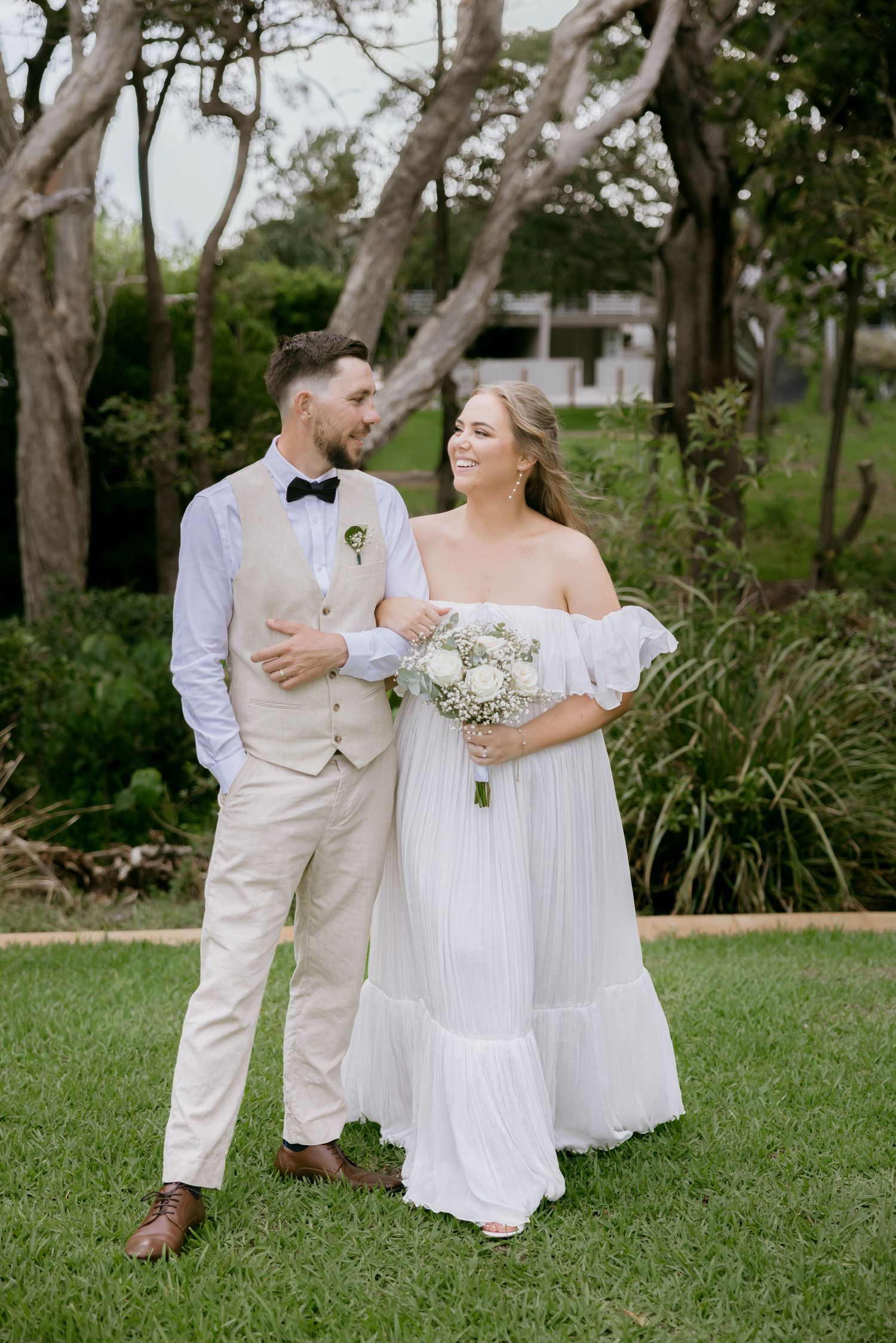 A couple in light-colored wedding attire share a romantic moment on a grassy lawn surrounded by trees.