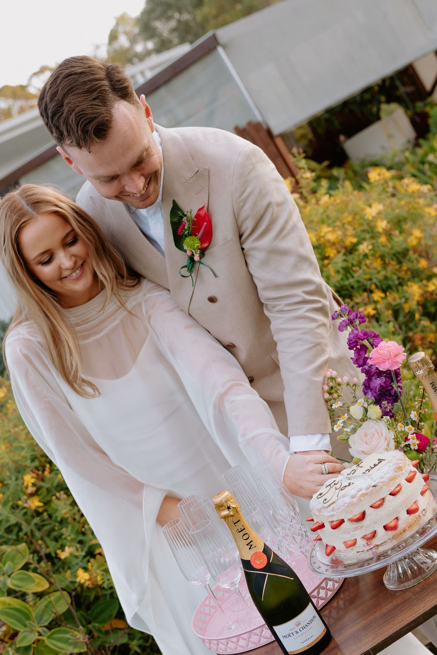 A couple cutting into a white wedding cake decorated with strawberries in a garden setting.