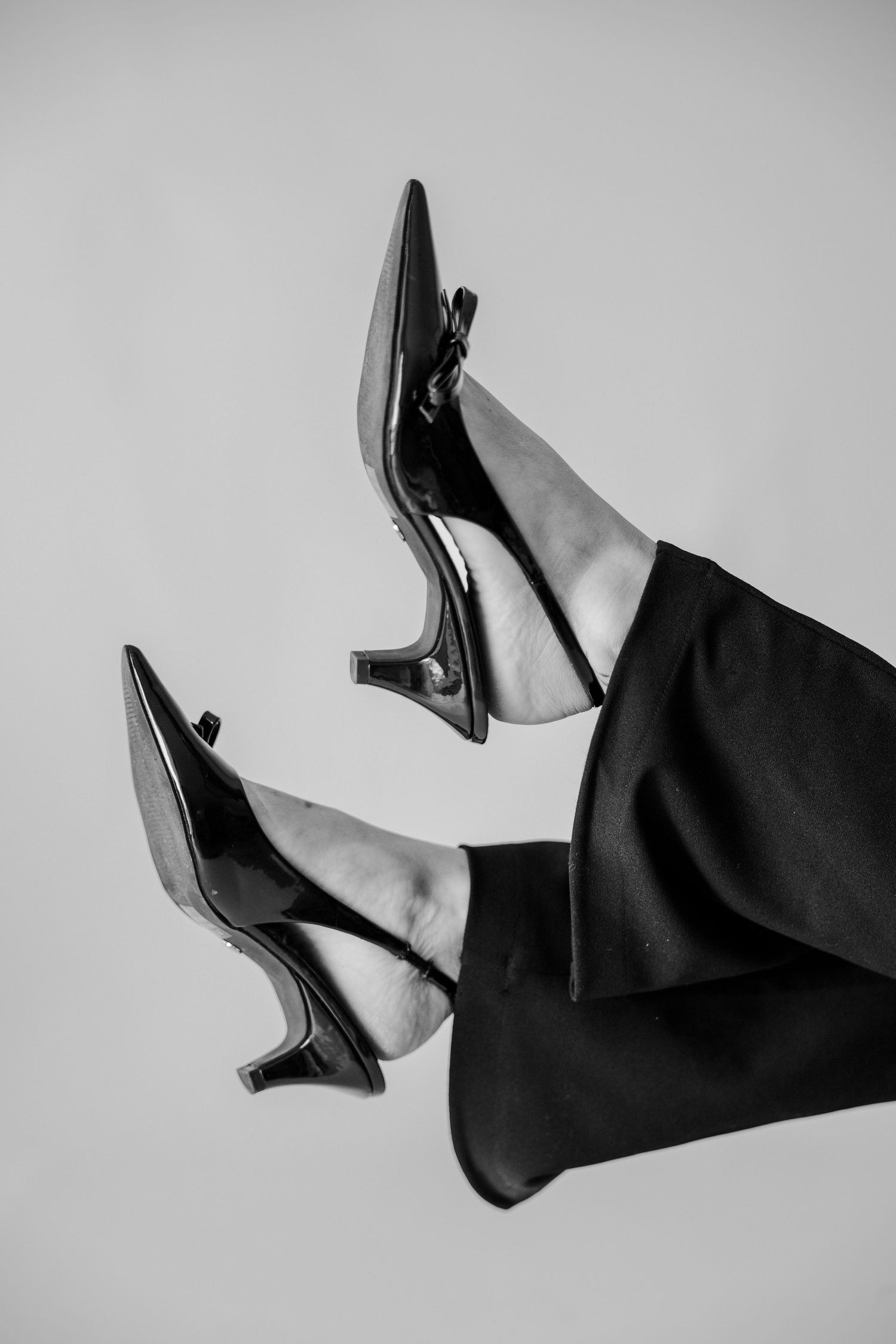 Black and white photo of womens legs raised in air wearing patent leather kitten heel slingback pumps and black trousers.