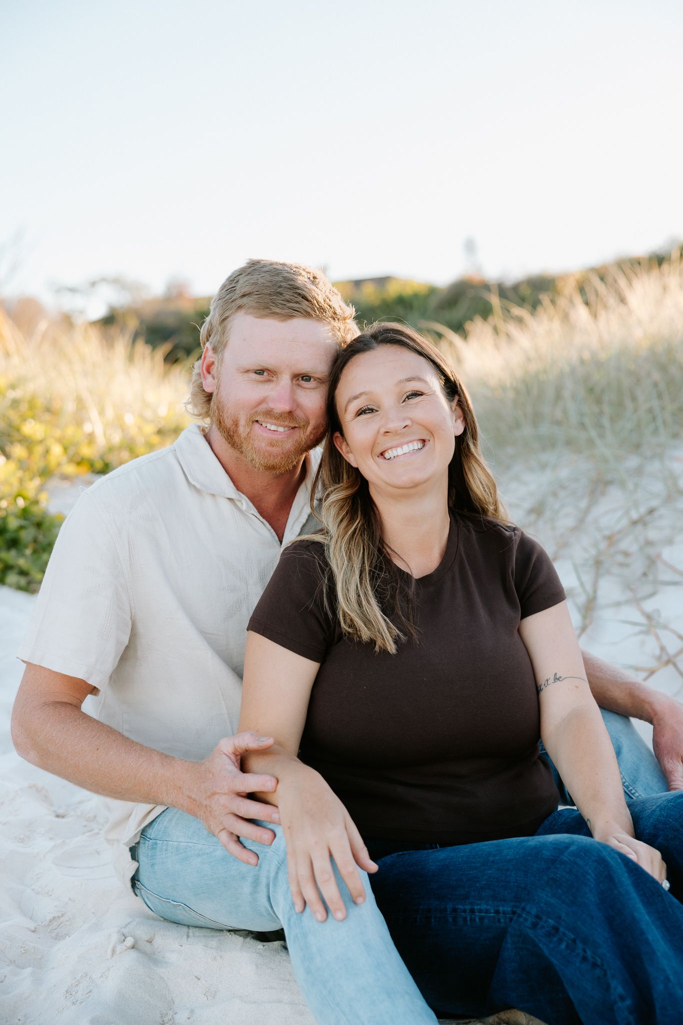 Smiling couple sitting together on white sand beach dunes, woman in brown top and man in beige shirt, sunny day.