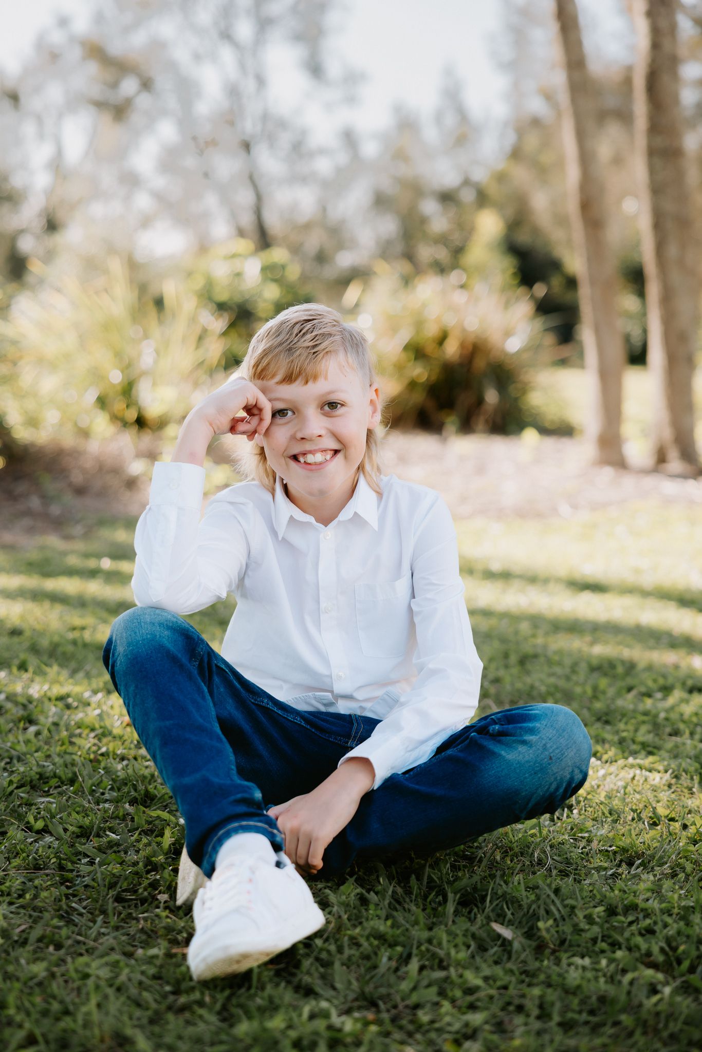 Smiling boy in white shirt and jeans sitting cross-legged on green grass in a sunny park.