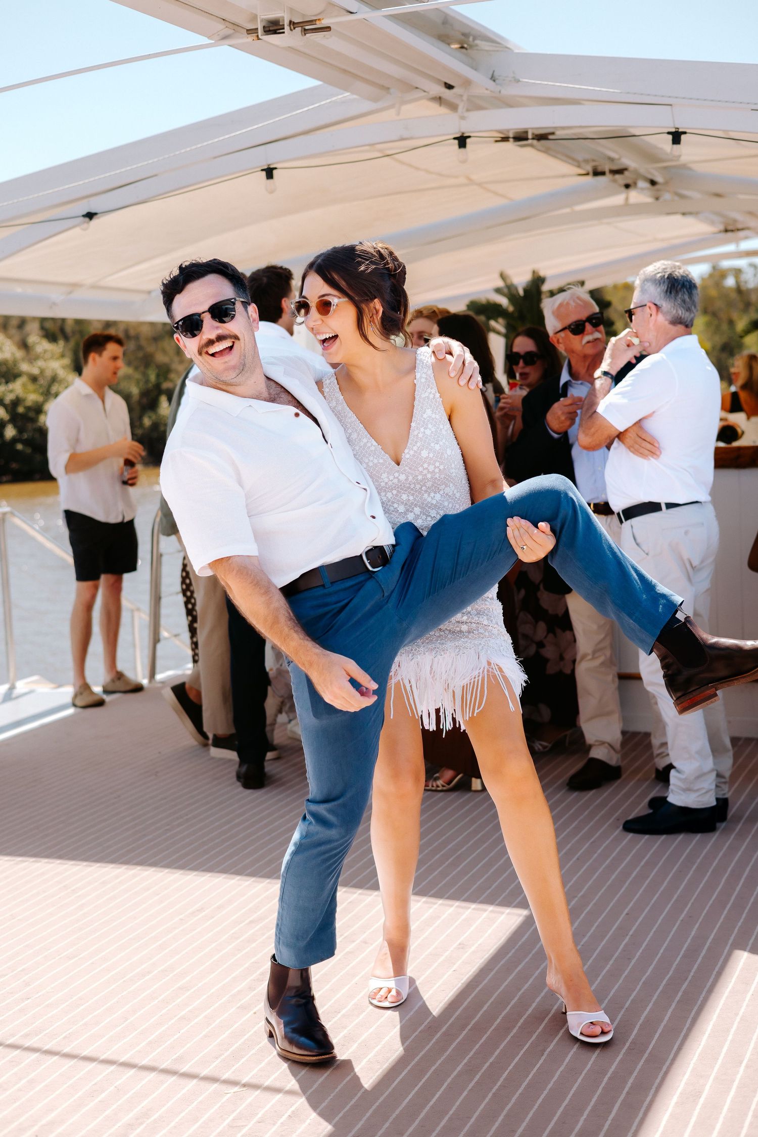 Couple dancing joyfully on a yacht deck, man lifting woman in white fringe dress, guests socializing in background.