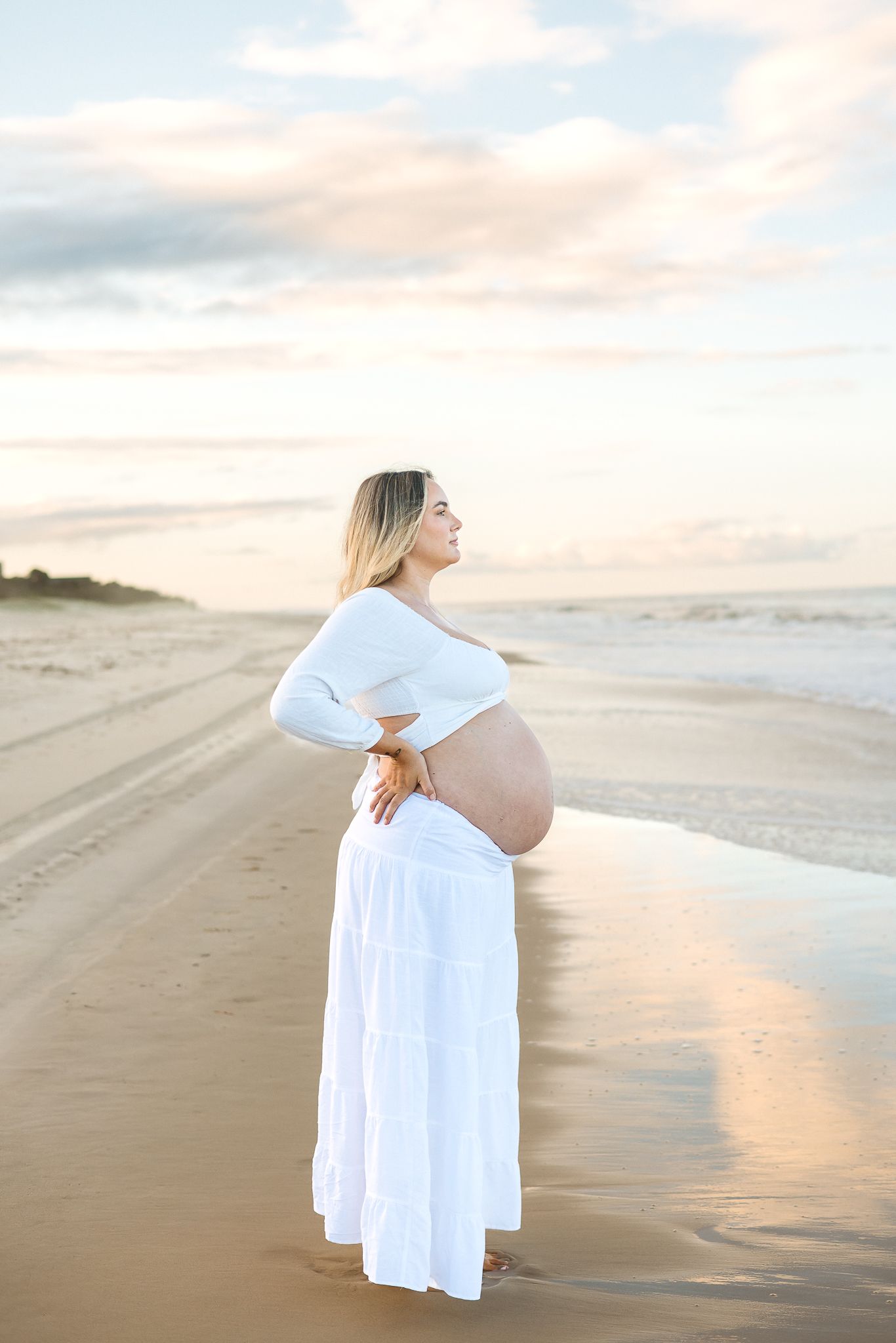 Pregnant woman in white crop top and skirt cradling baby bump on a serene beach at sunset.