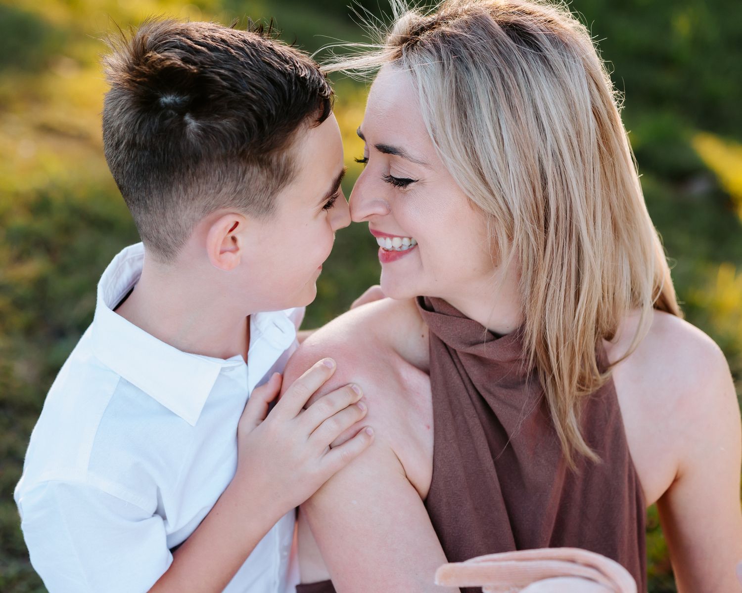 A smiling woman and young boy share a tender nose-to-nose moment outdoors in warm natural light.