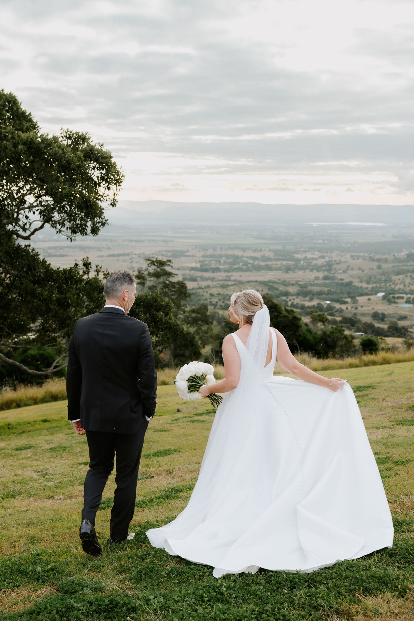Bride and groom walk hand in hand on a grassy hilltop with sweeping valley views on their wedding day.