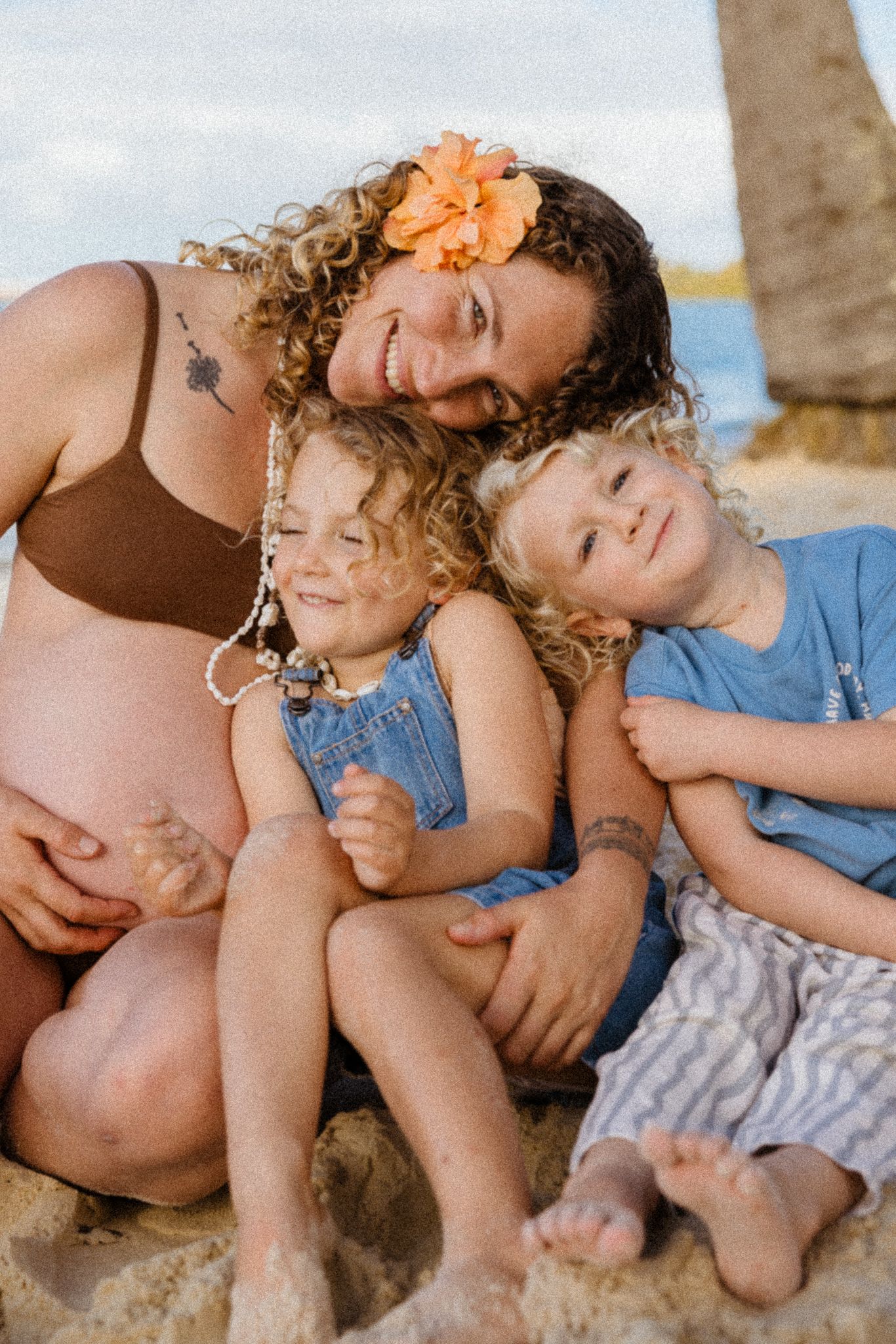 Pregnant woman with flower in curly hair smiles with two young curly-haired children on a sandy beach.