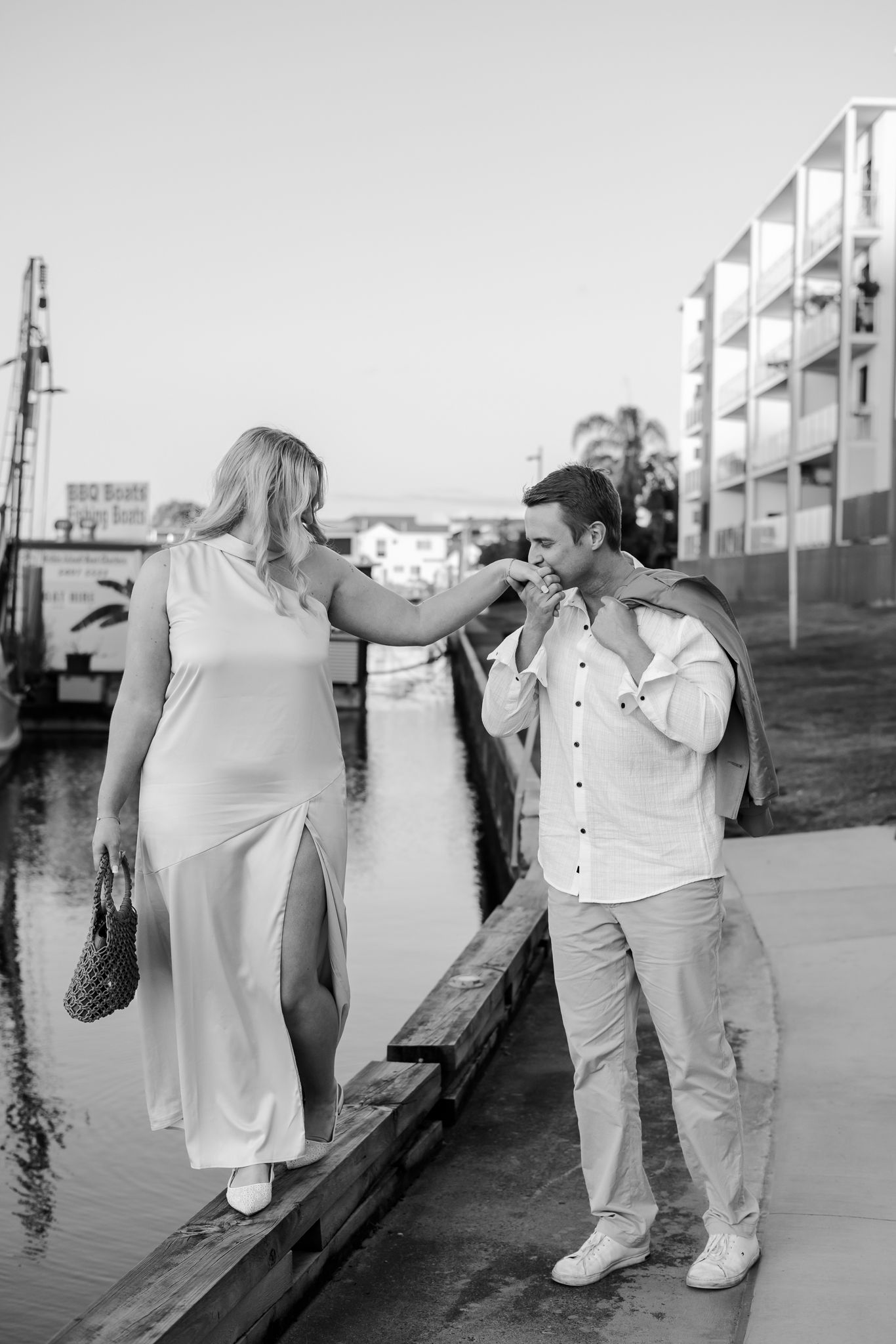 Black and white photo of a couple on a dock, man kissing woman's hand near a waterfront with buildings in background.