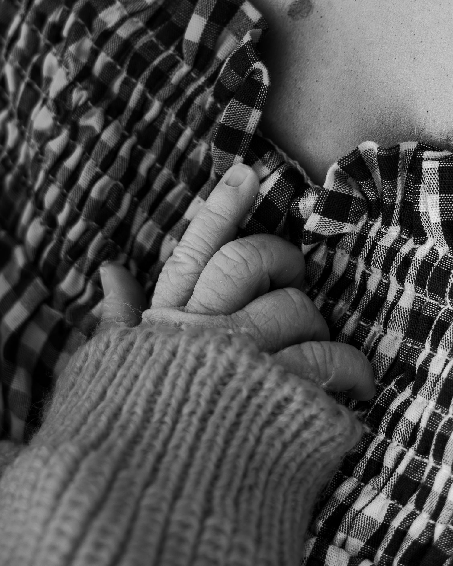Black and white photo of an adult hand gently holding a newborn baby's tiny hand, wearing plaid clothing.