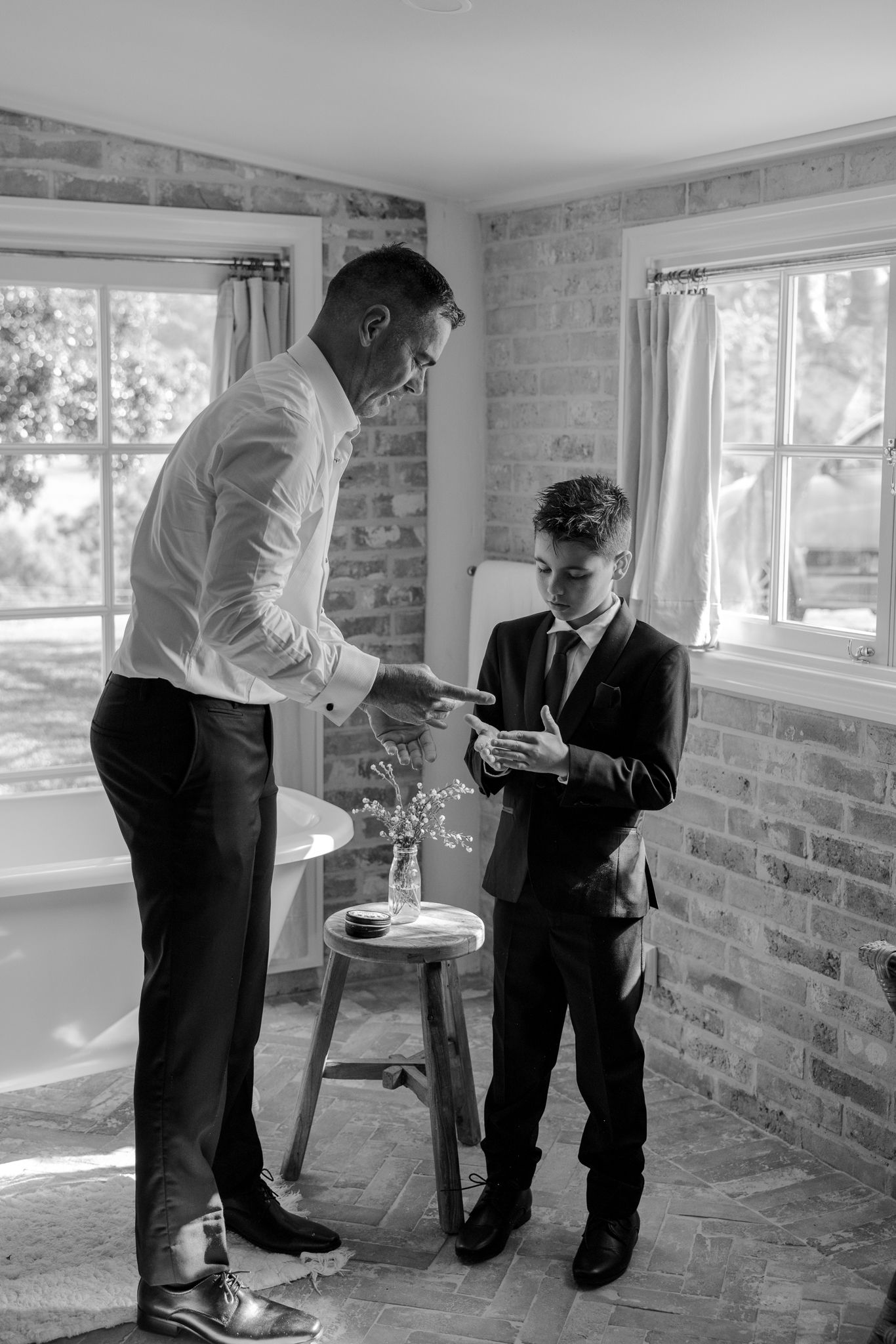 Black and white photo of groom and young boy in suits preparing for a wedding indoors.