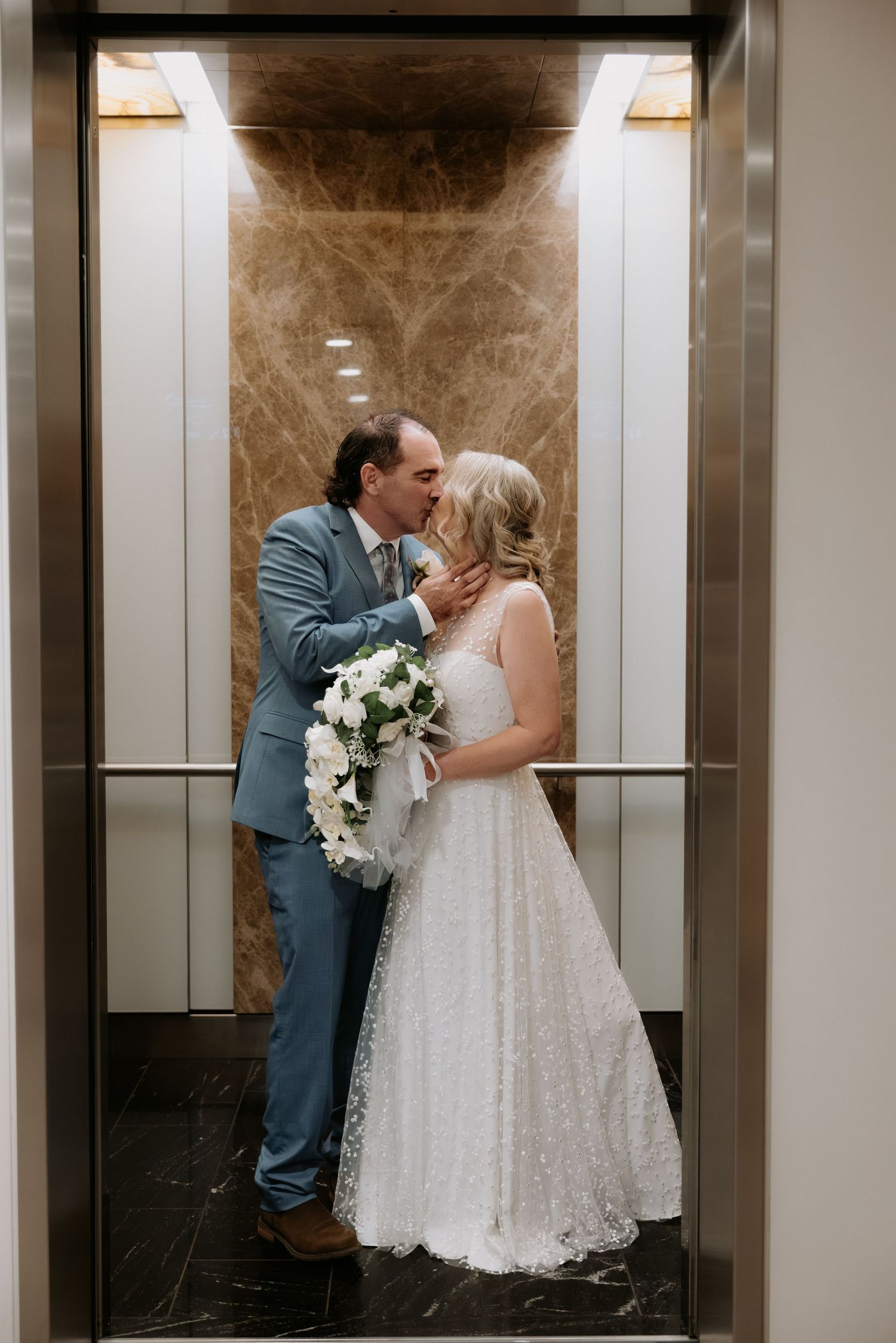 A romantic wedding couple shares intimate kisses in an elegant elevator with marble walls and white floral bouquet.