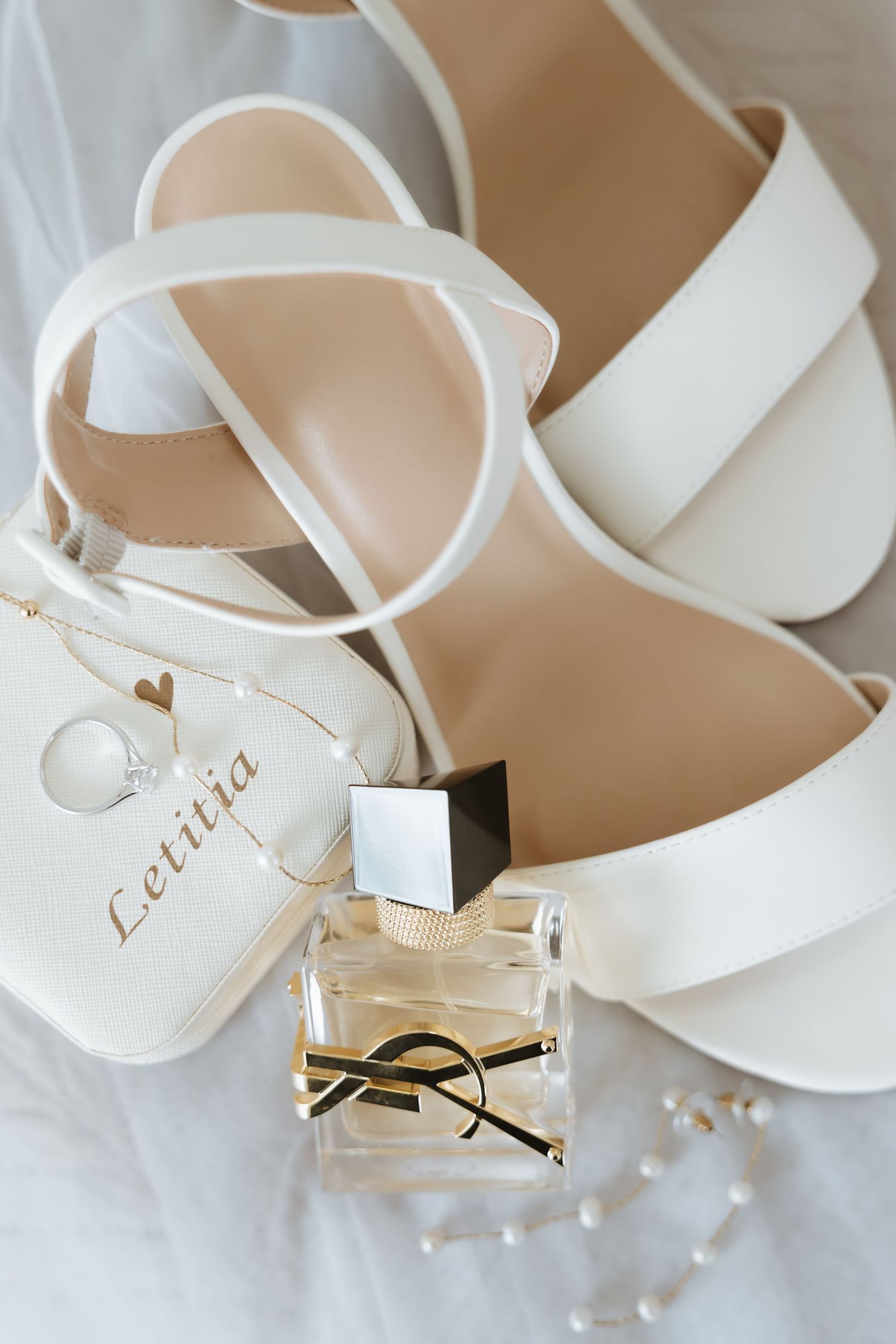 White bridal sandals with straps arranged with YSL perfume bottle and small pearls on light background.