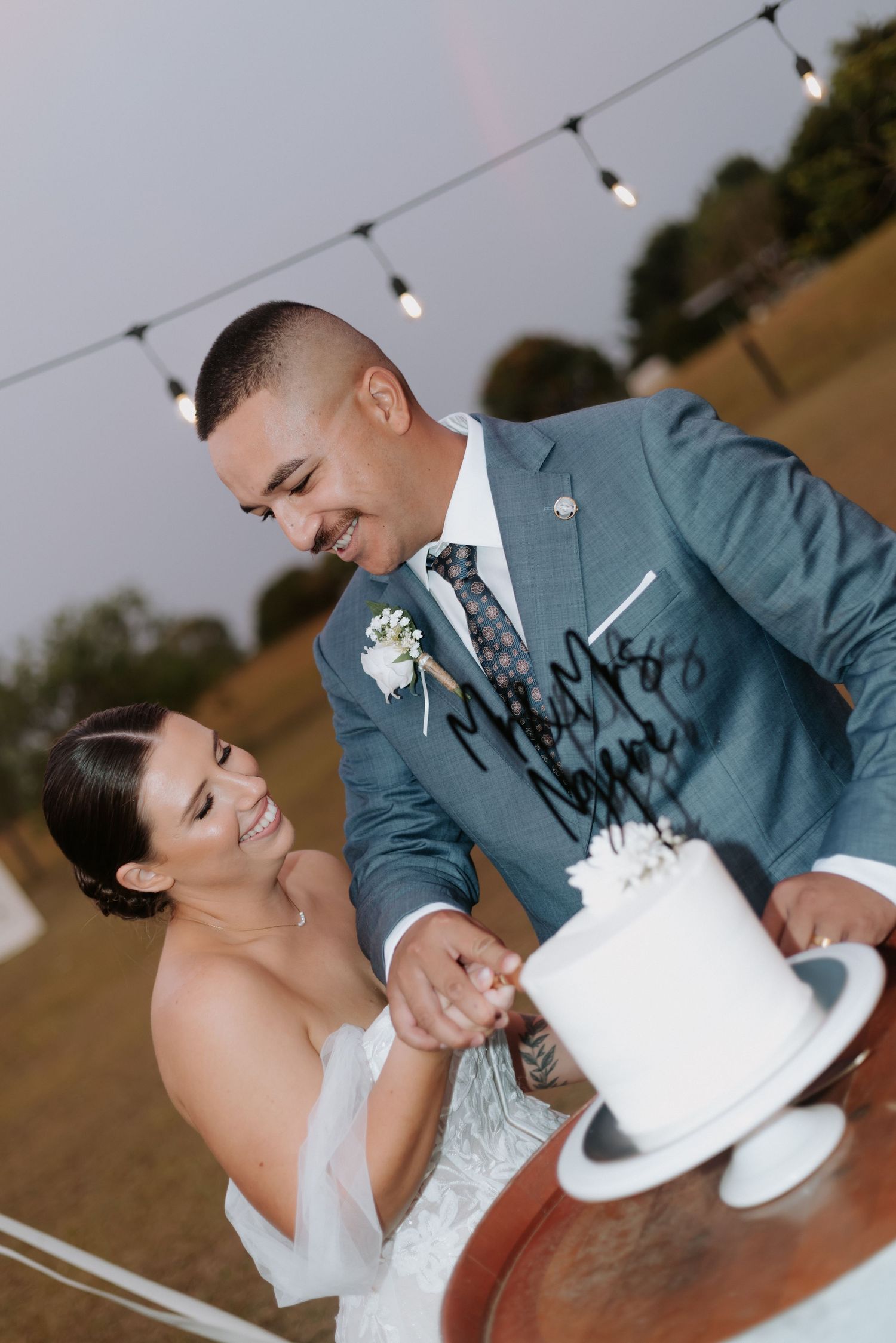Bride and groom cut their white wedding cake together under string lights at dusk.