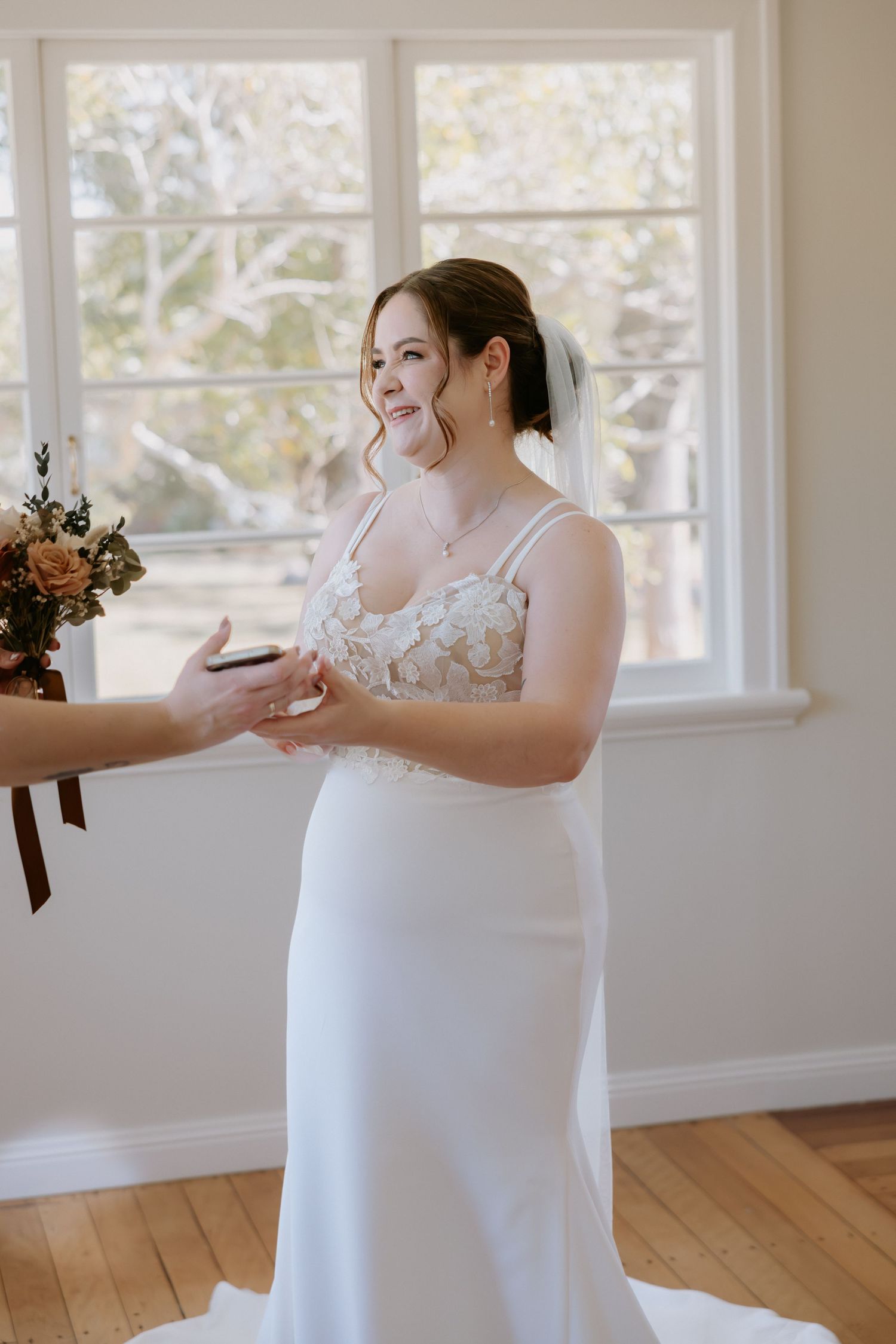 Bride in a white lace dress standing in a bright room with large windows.
