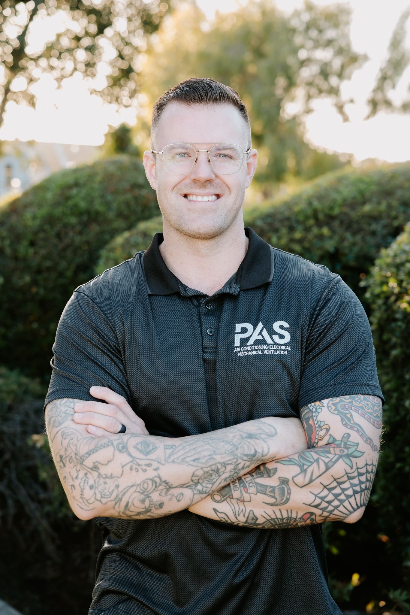 Smiling man with tattoos in a black PAS polo shirt standing with arms crossed outdoors near green hedges.