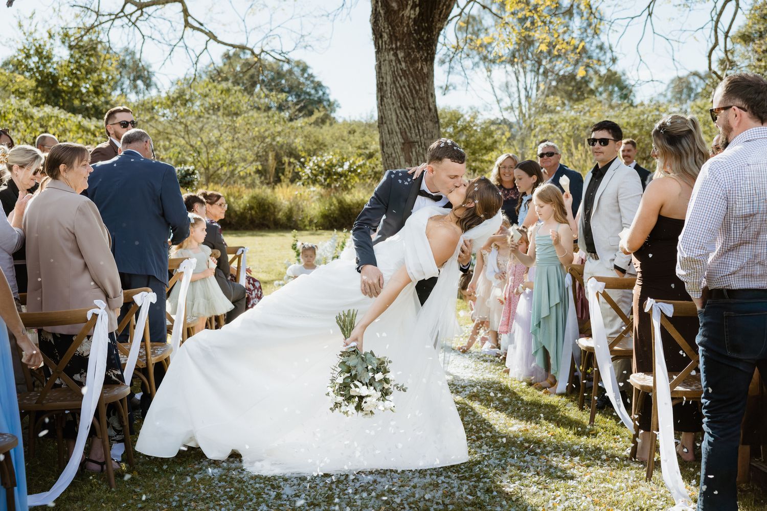 Bride and groom share a romantic dip kiss at outdoor wedding ceremony aisle lined with guests and flower girls.