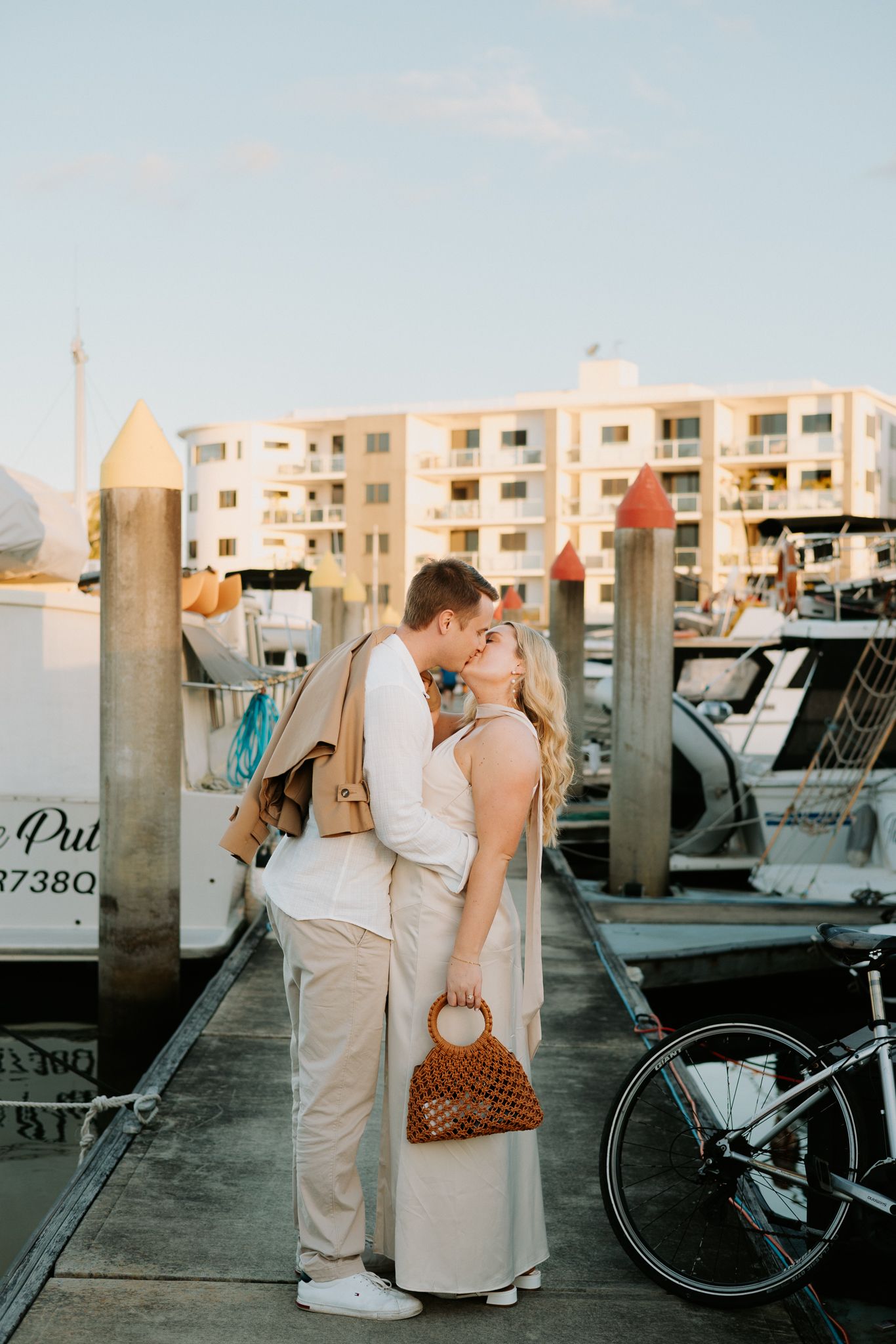 Couple kissing on a marina dock at golden hour, with boats and waterfront apartments in the background.