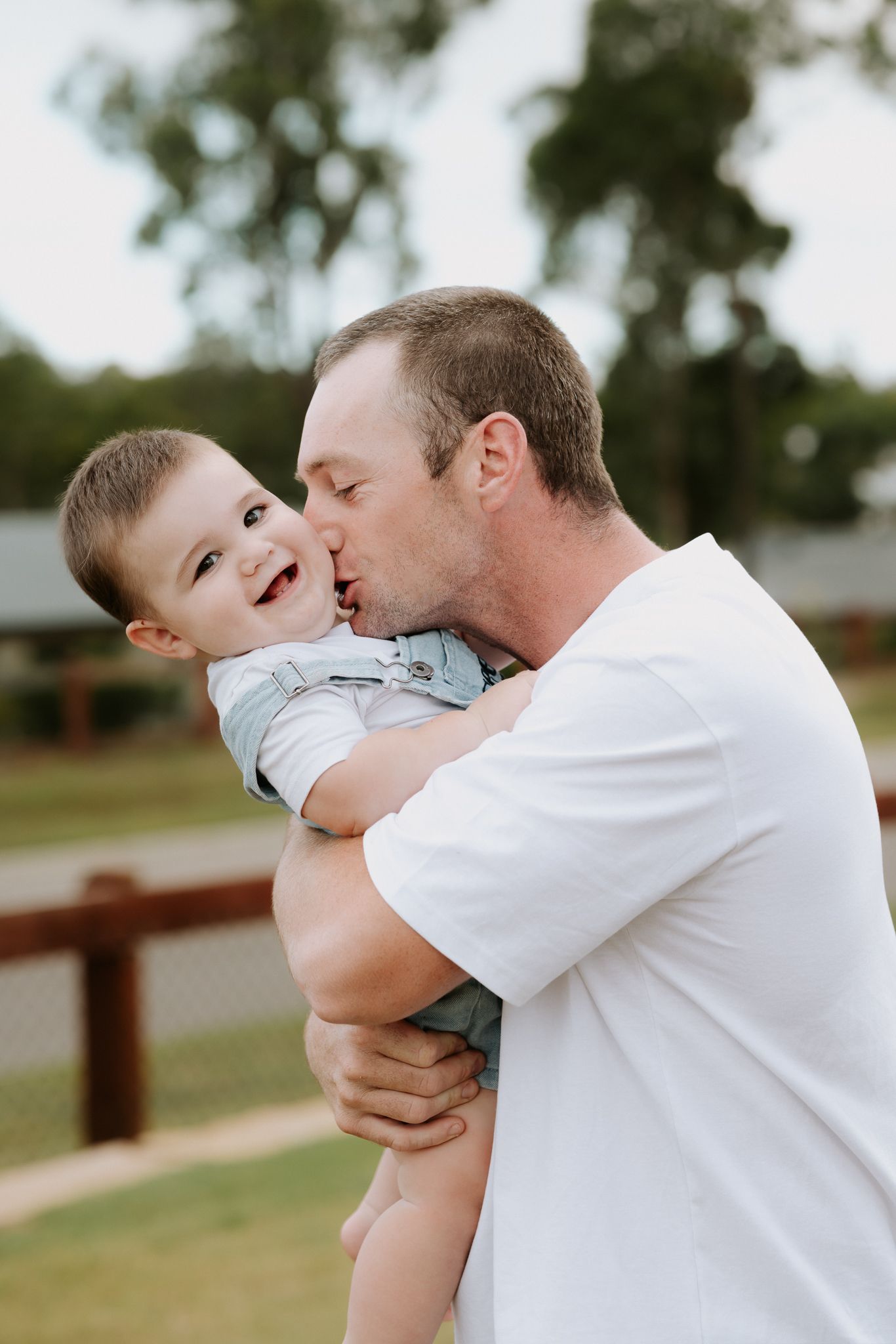 A father kisses his laughing baby boy while holding him outdoors near a wooden fence.