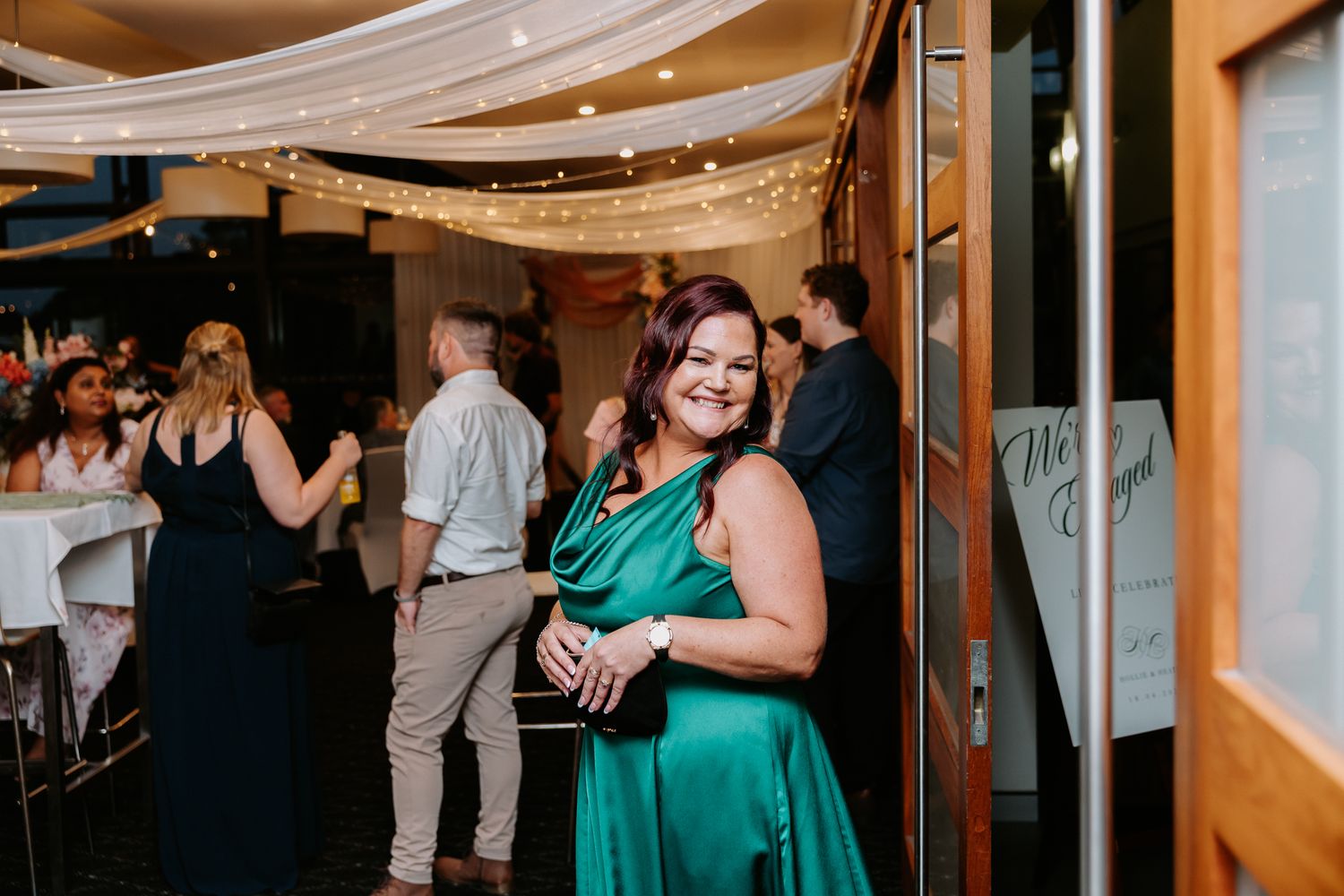 Woman in elegant green satin dress smiling at wedding reception with fairy lights and guests in background.