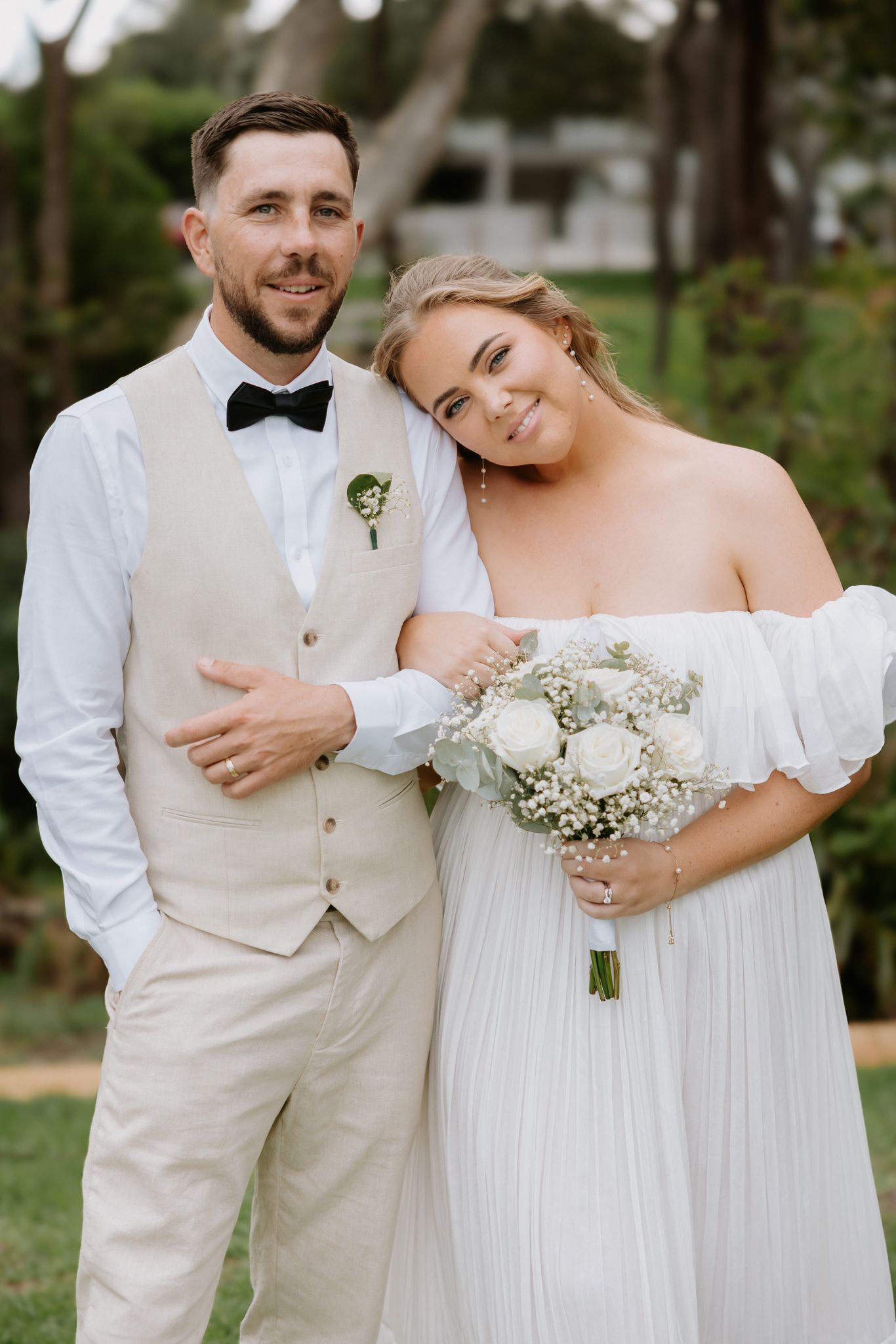 Smiling bride and groom posing outdoors, bride holding white bouquet, groom wearing beige vest and bow tie.