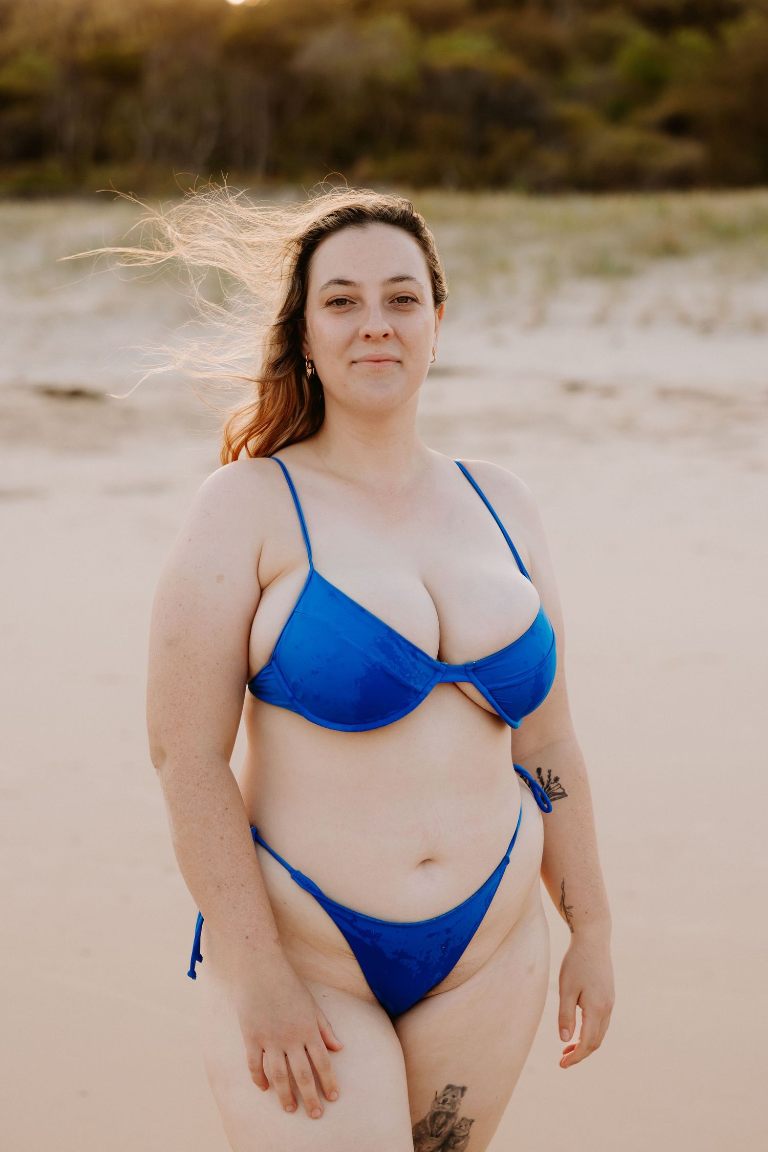 Woman in blue bikini standing on sandy beach with wind-blown hair and tattoos, golden hour lighting.