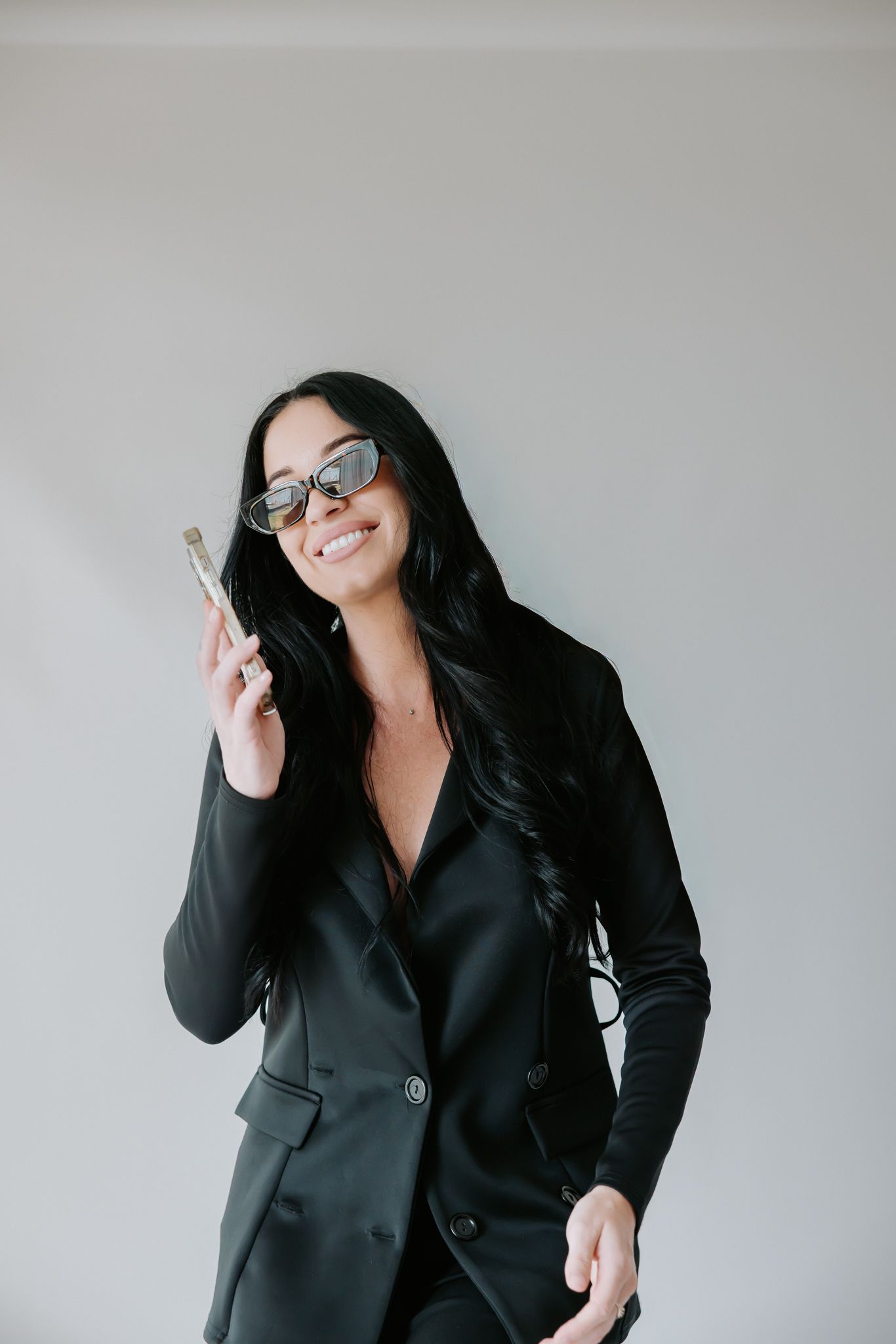 Smiling woman in black suit holding a paintbrush, wearing sunglasses, posing against a light gray background.