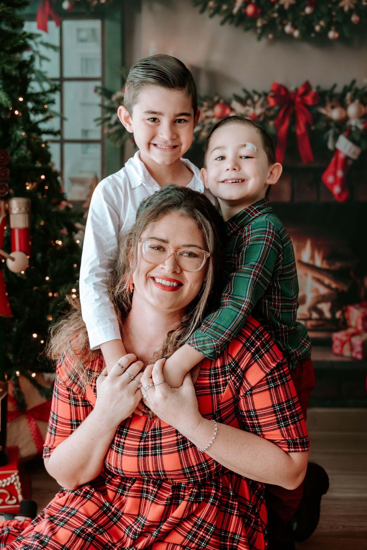 A smiling mother in red plaid dress poses with two happy boys near a decorated Christmas tree and fireplace.