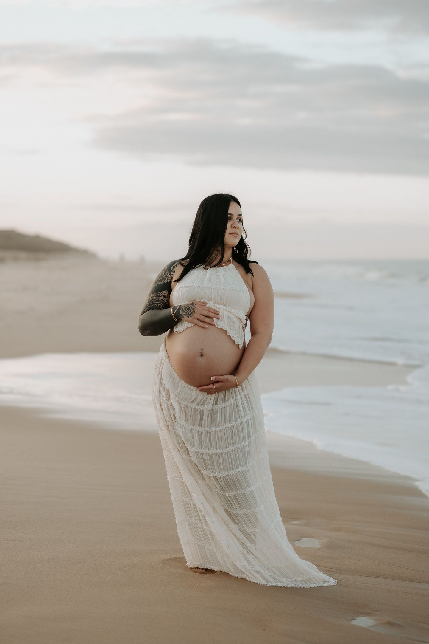 A pregnant woman in a flowing white dress poses on a misty beach at sunset.