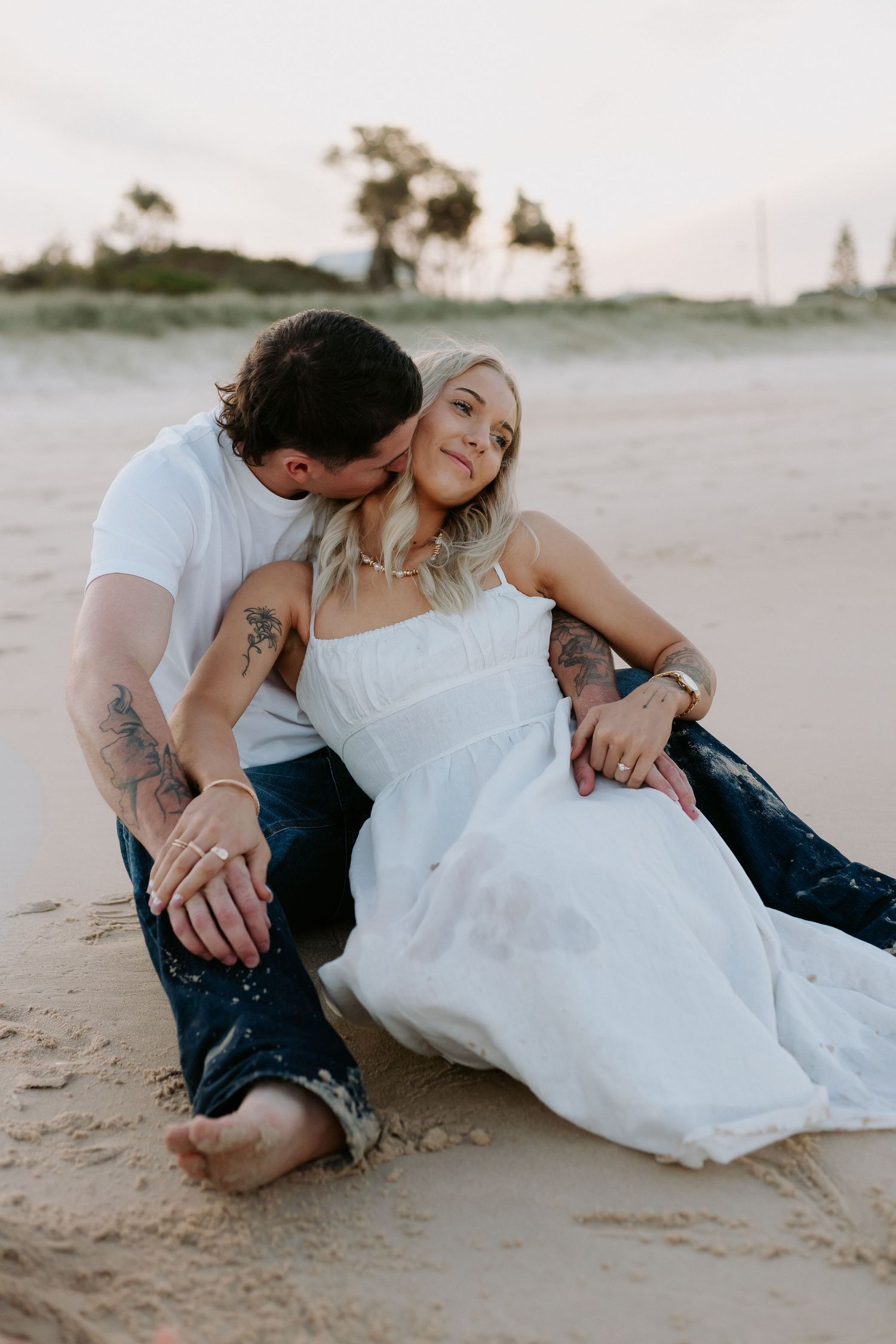 Couple sitting on beach sand, man kissing woman's cheek, both with tattoos, woman in white dress at sunset.