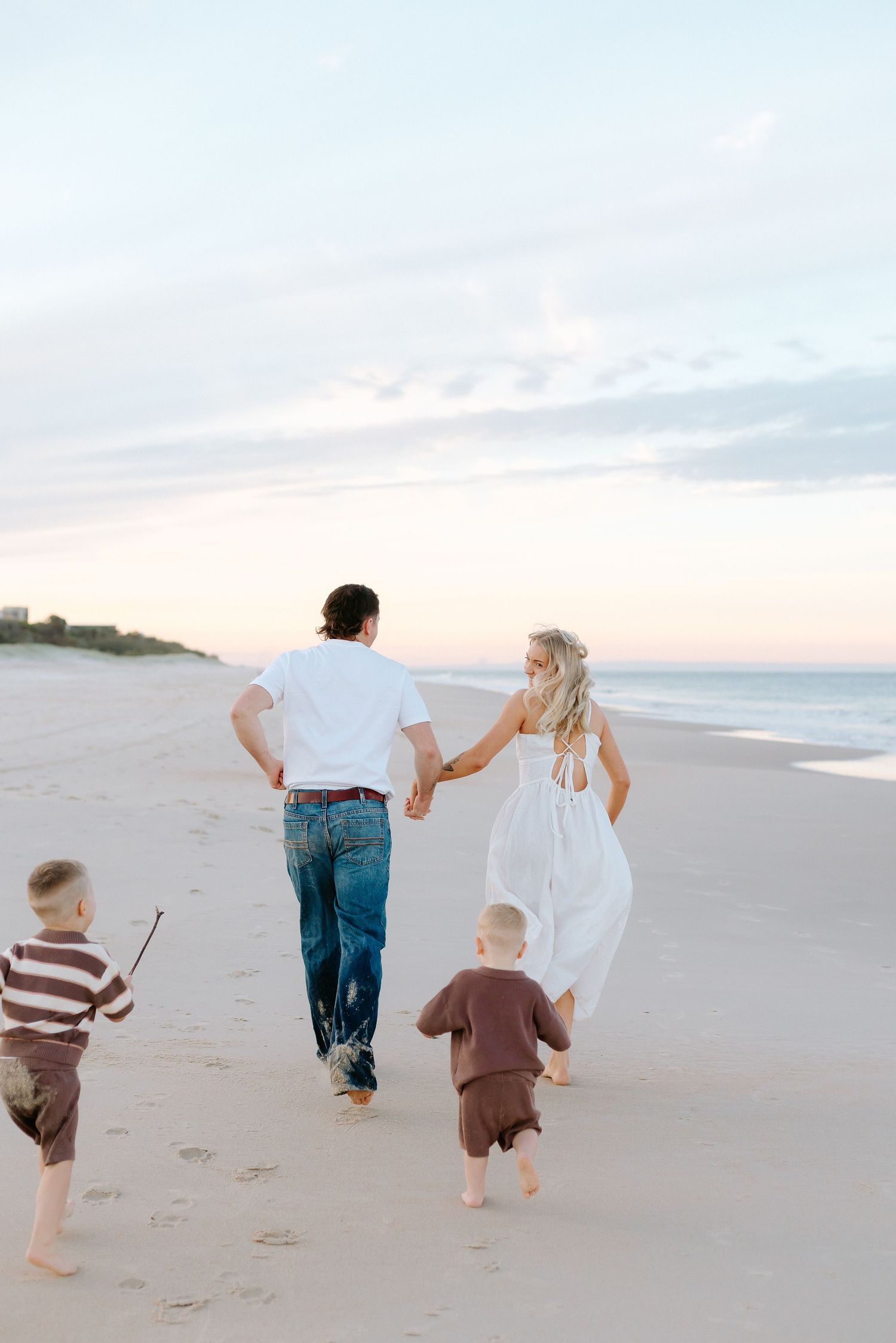 Family of four walking hand-in-hand on a sandy beach at sunset, two young boys running alongside their parents.