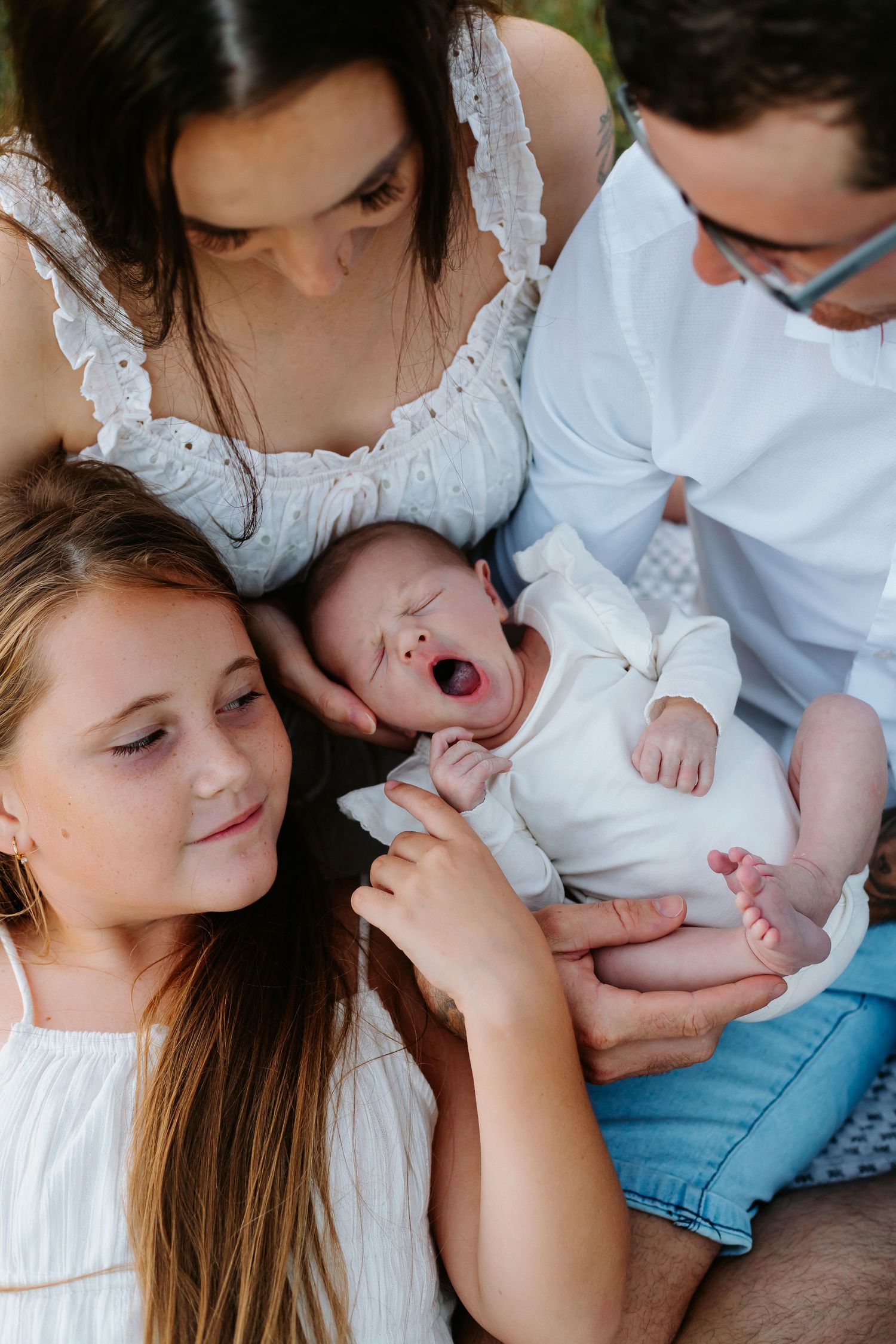 Family gathered around yawning newborn baby, older sister and parents in white outfits sharing a tender moment outdoors.