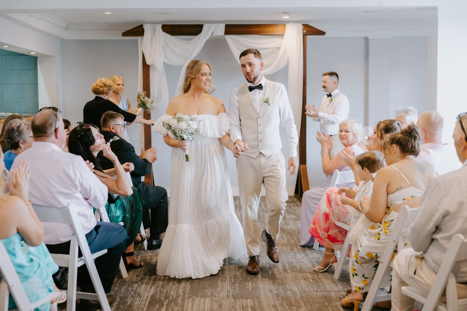 Bride and groom walk down the aisle together after wedding ceremony, guests applauding in decorated indoor venue.