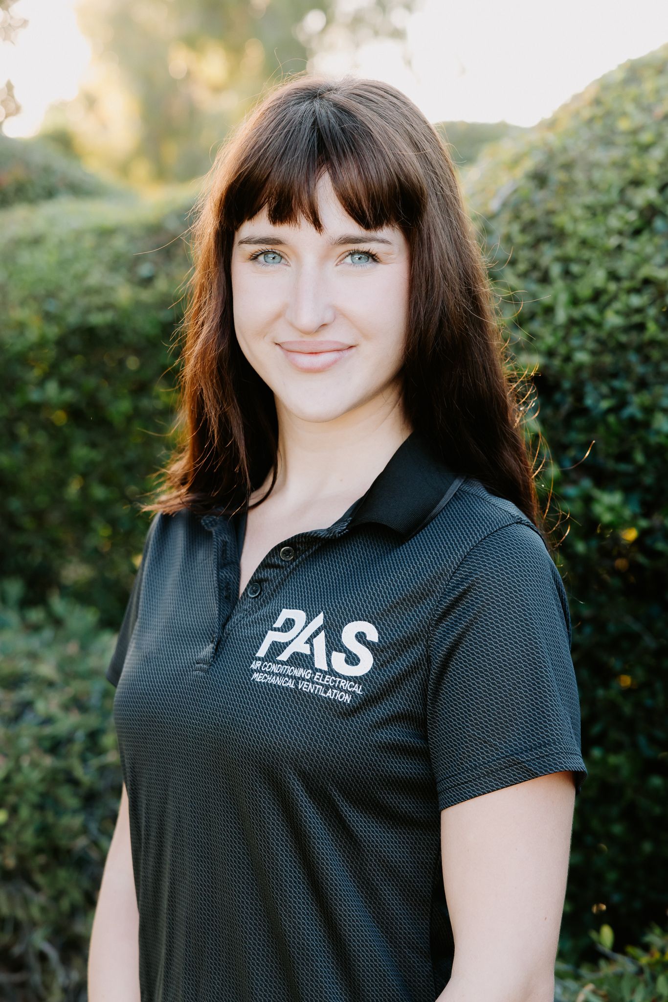 Woman with dark hair wearing a PAS Air Conditioning Electrical Mechanical Ventilation polo shirt outdoors.